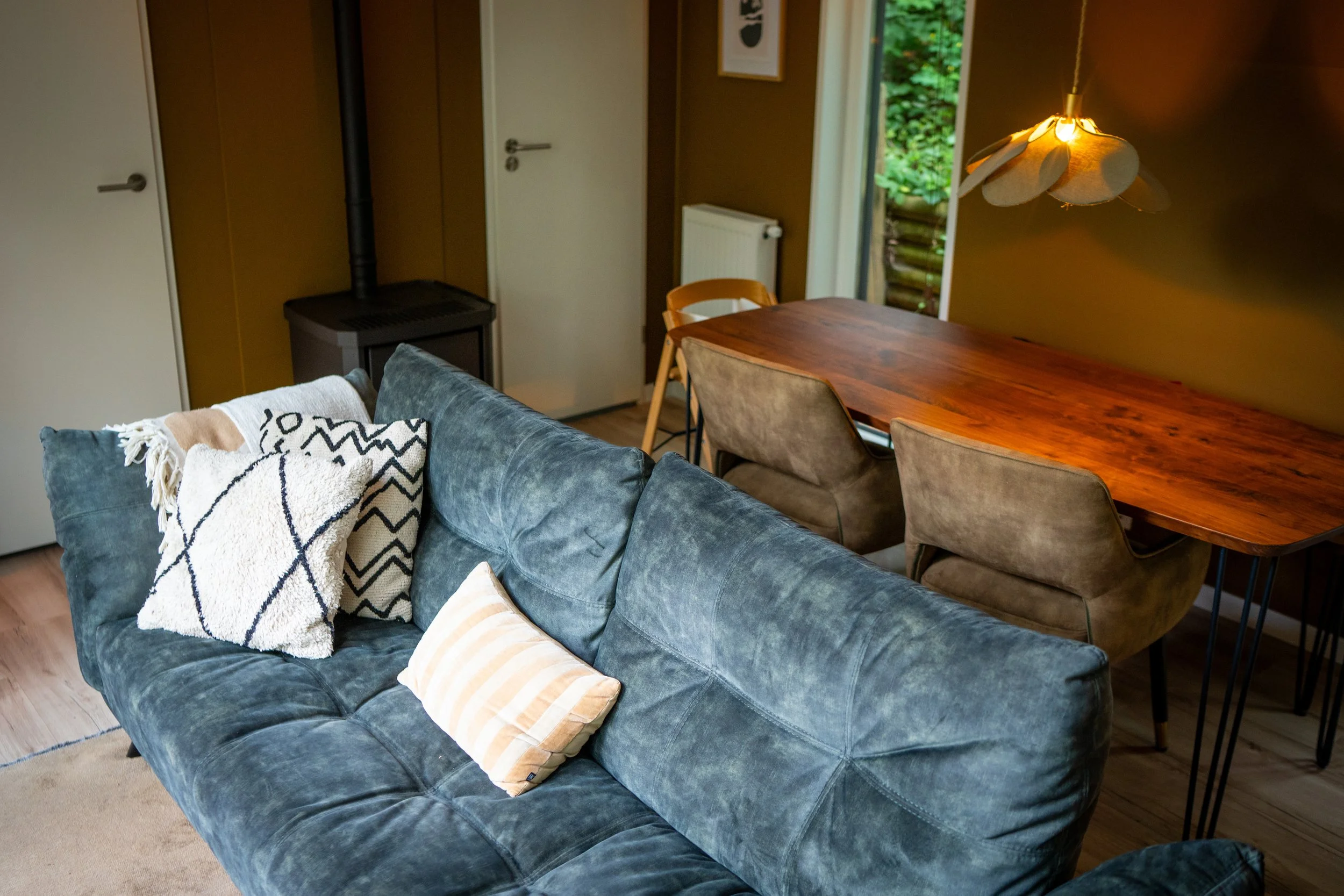 Interieur van een woonkamer met een blauwe bank met decoratieve kussens, een houten eettafel, stoel en hanglamp, met uitzicht op een tuin via een open deur.