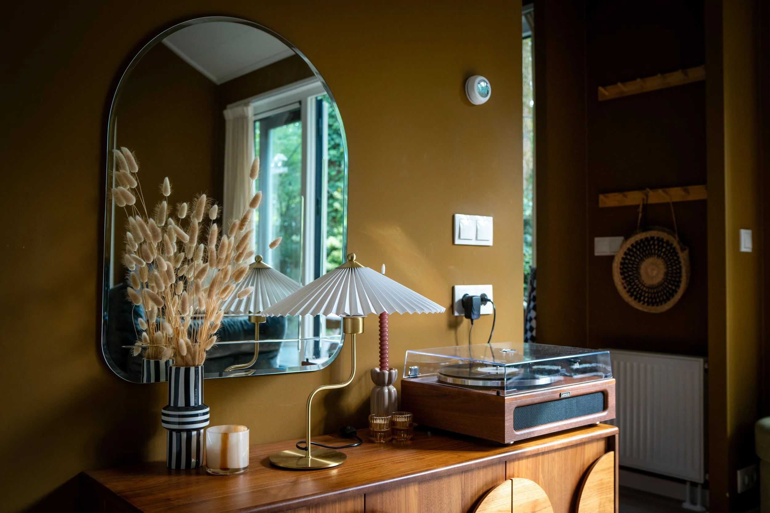 Een interieur met een ronde spiegel, een vaas met gedroogde bloemen, een tafelklok, een platenspeler en een lamp op een houten meubel, met een raam en een muur in warme tinten.