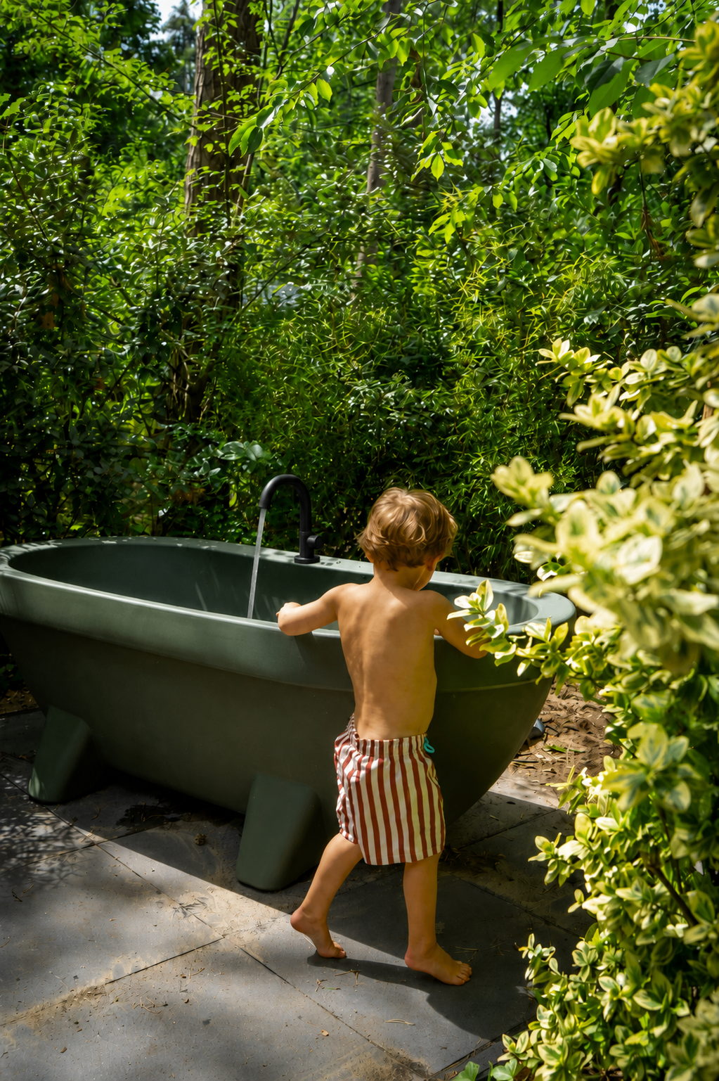 Kleine jongen met gestripeshortjes wast zich in een buitensas, omgeven door groene struiken en bomen in de tuin.