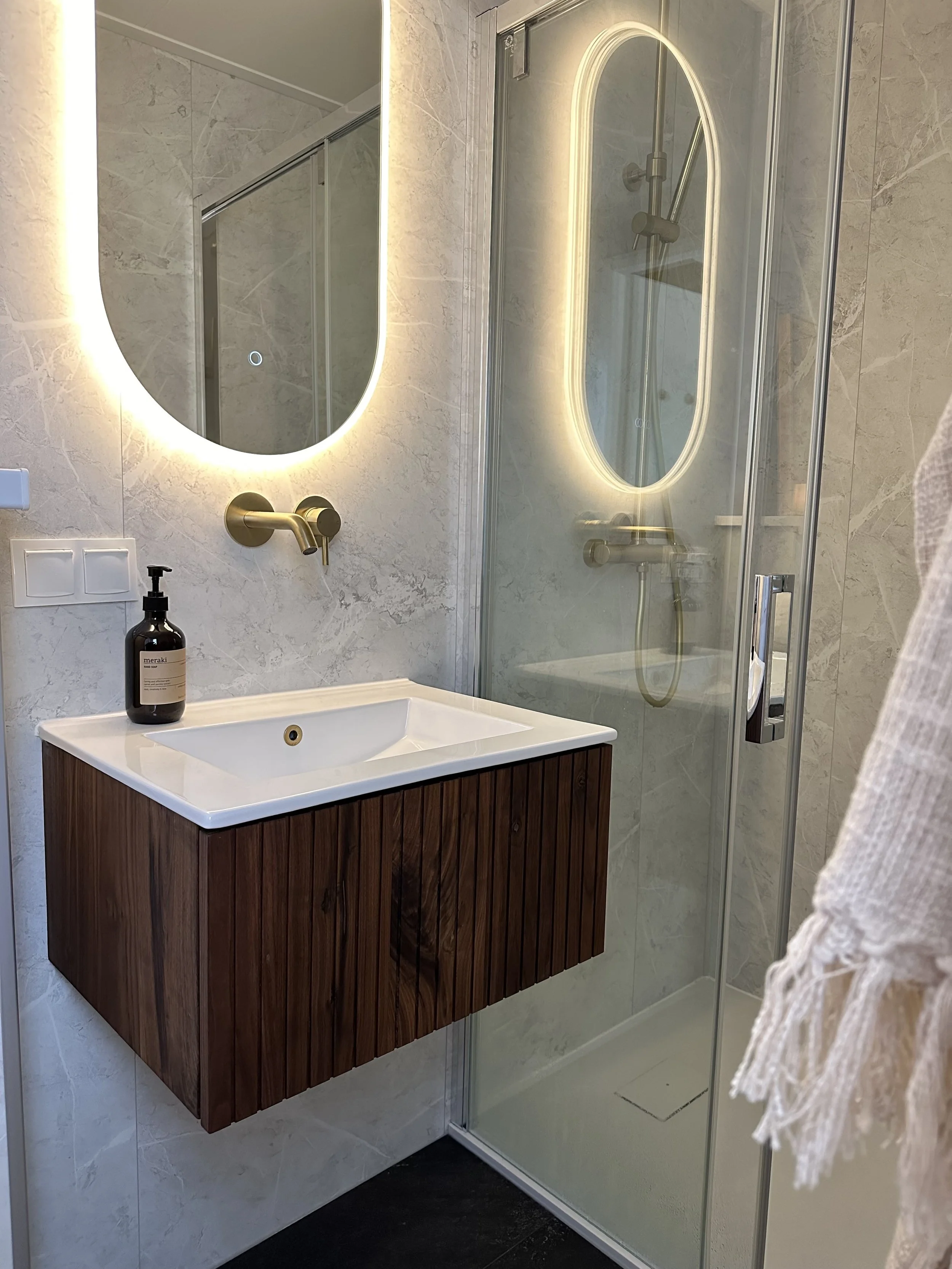 Moderne badkamer met een spiegel met ingebouwd licht, een houten wastafelonderkast, een zwarte zeeppomp, een spiegelkast en een inloopdouche met glazen deuren en een regendouchekop.