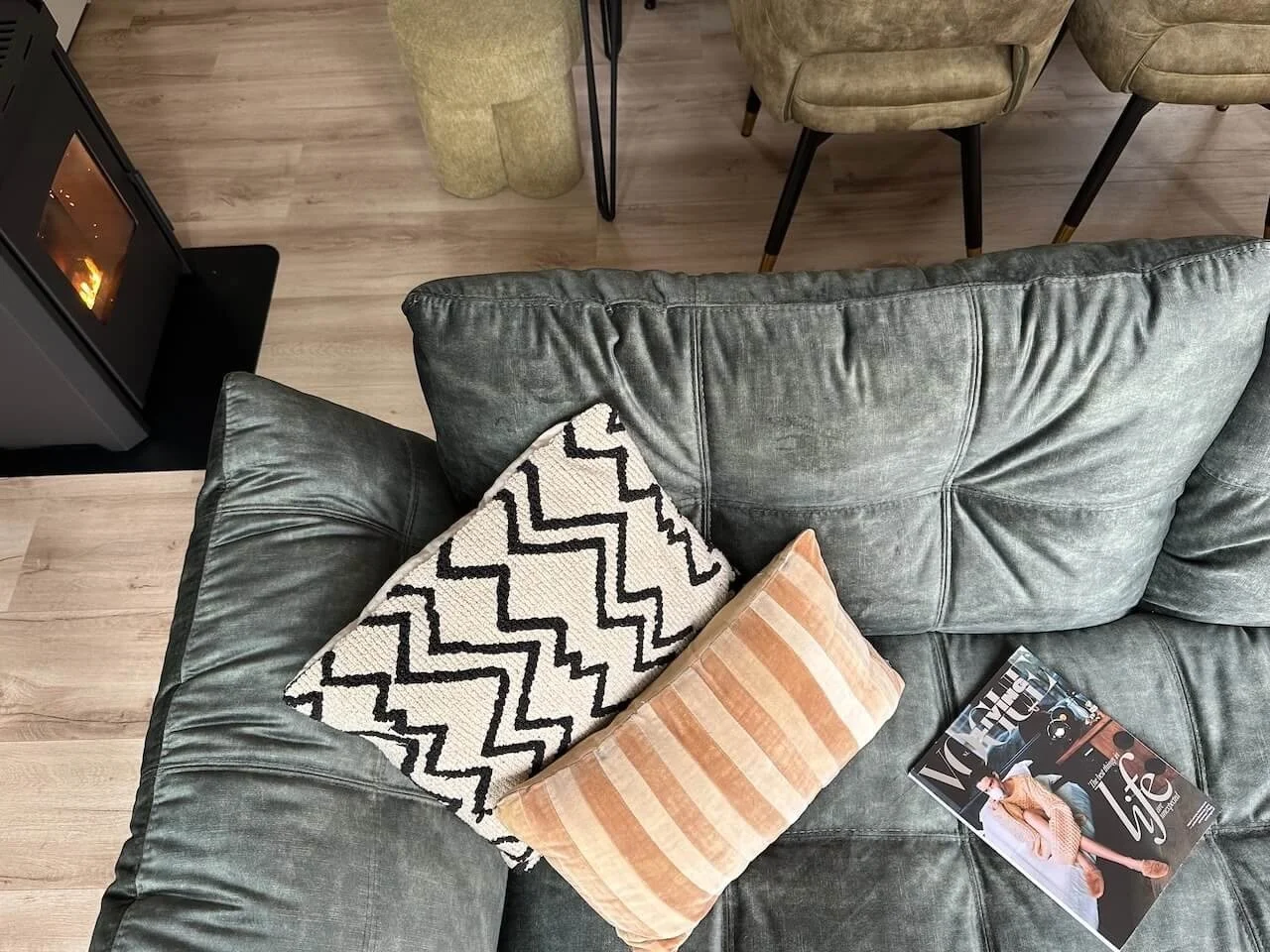 Een groene leren bank met twee kussens, een met zwart-witte zigzagpatronen en een met lichtoranje strepen, en een tijdschrift, in een woonkamer met houten vloer en een houtkachel.