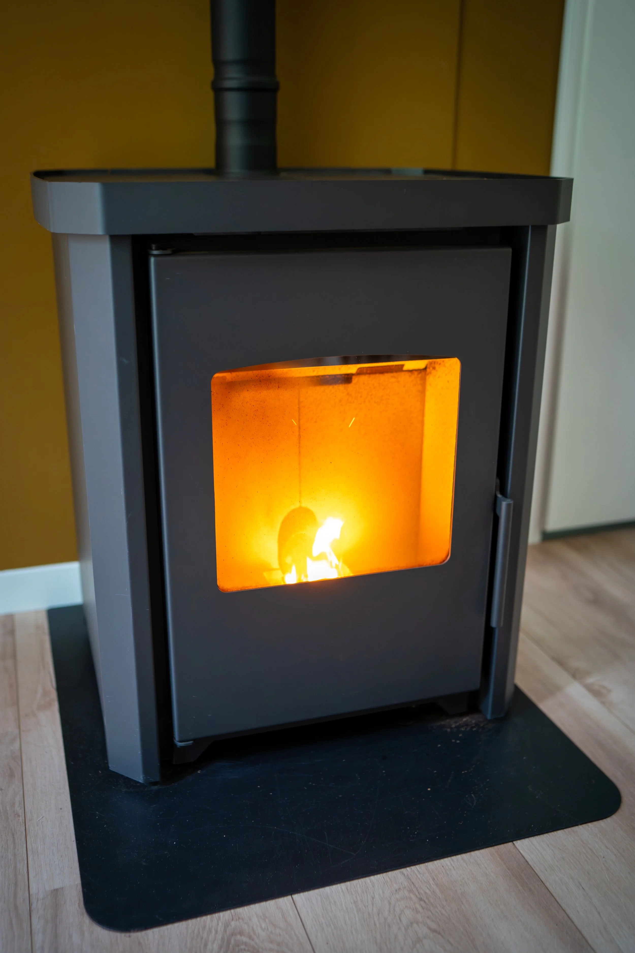 Elektrische kachel met een open deurtje, brandend binnenin, geplaatst op een zwarte mat op een houten vloer.