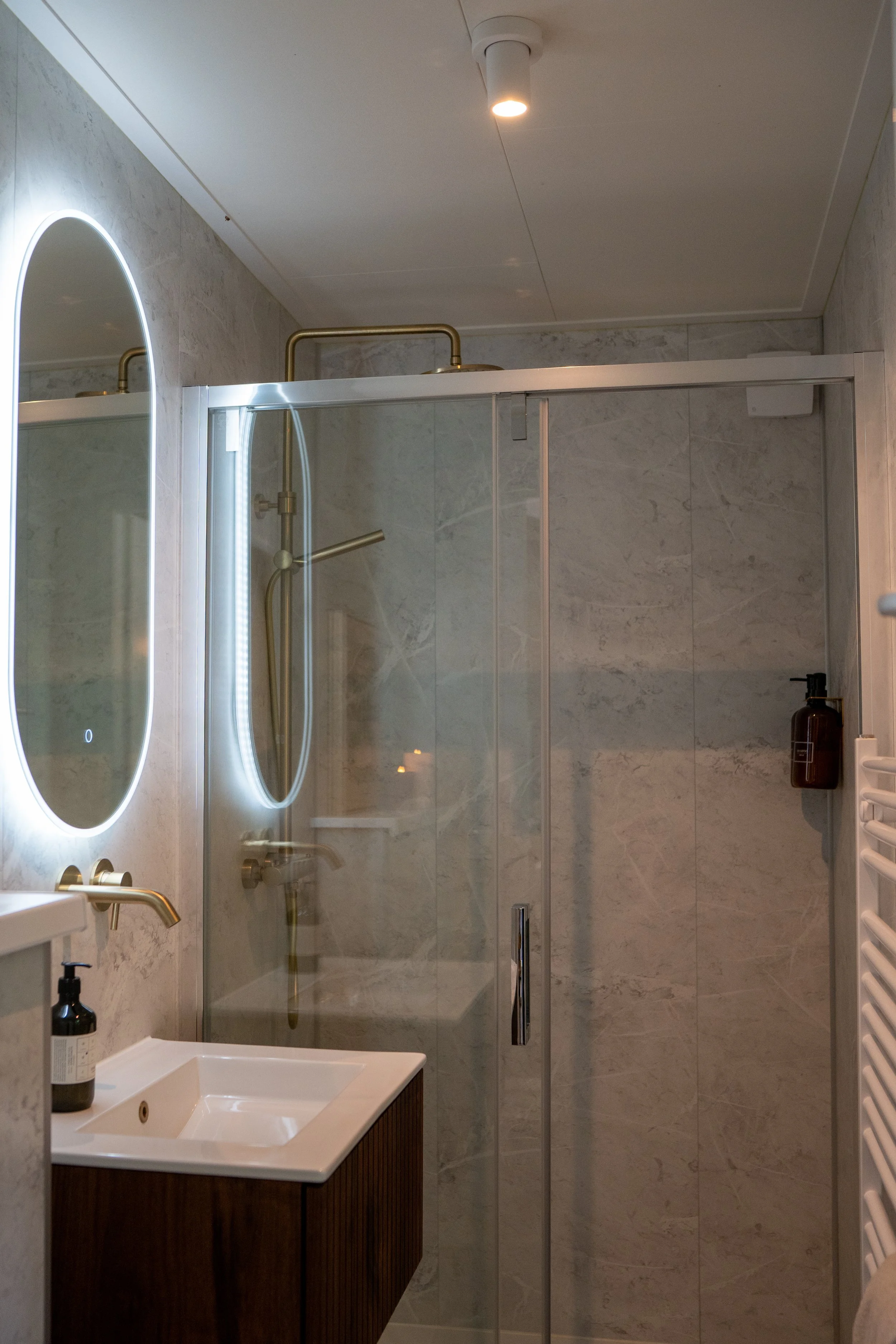 Een moderne badkamer met een dwarsdeurtruimte schort, een spiegel met led-verlichting, een kleine wastafel met kraan en een fles zeep op de rand, en een muur met marmerlook tegels.
