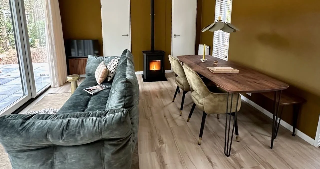 Interieur van een woonkamer met een groene bank, een houten eettafel met vier stoelen, een houtkachel, en grote ramen die naar een tuin leiden.