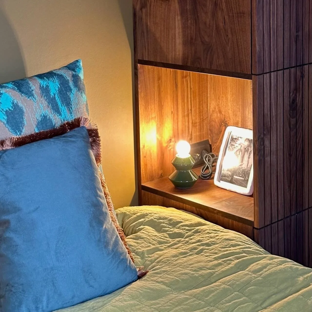 Een slaapkamer met een bed dat een geeldekbed en blauwe kussens heeft, en een houten nachtkastje met een lamp, een foto in een digitale fotolijst en een stekkerdoos.