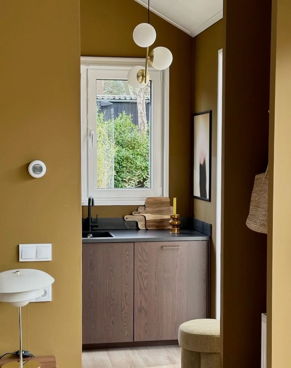 Een kleine keuken met een zwart werkblad, houten kasten, een raam met groen uitzicht, en muurdecoraties.