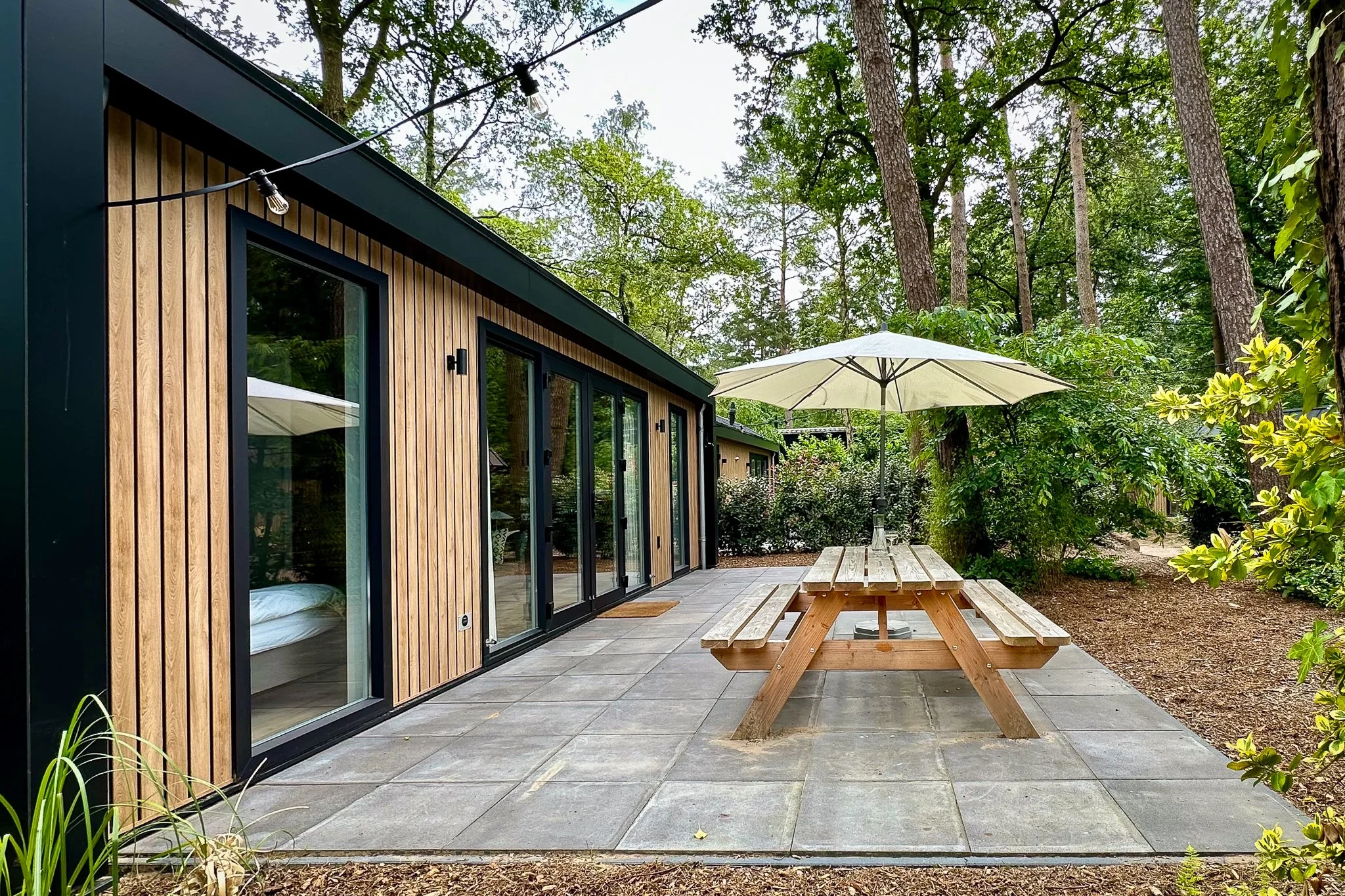 Een moderne houten woning met grote glazen deuren, een patio met een picknicktafel en een grote parasol, omgeven door groene bomen en struiken.