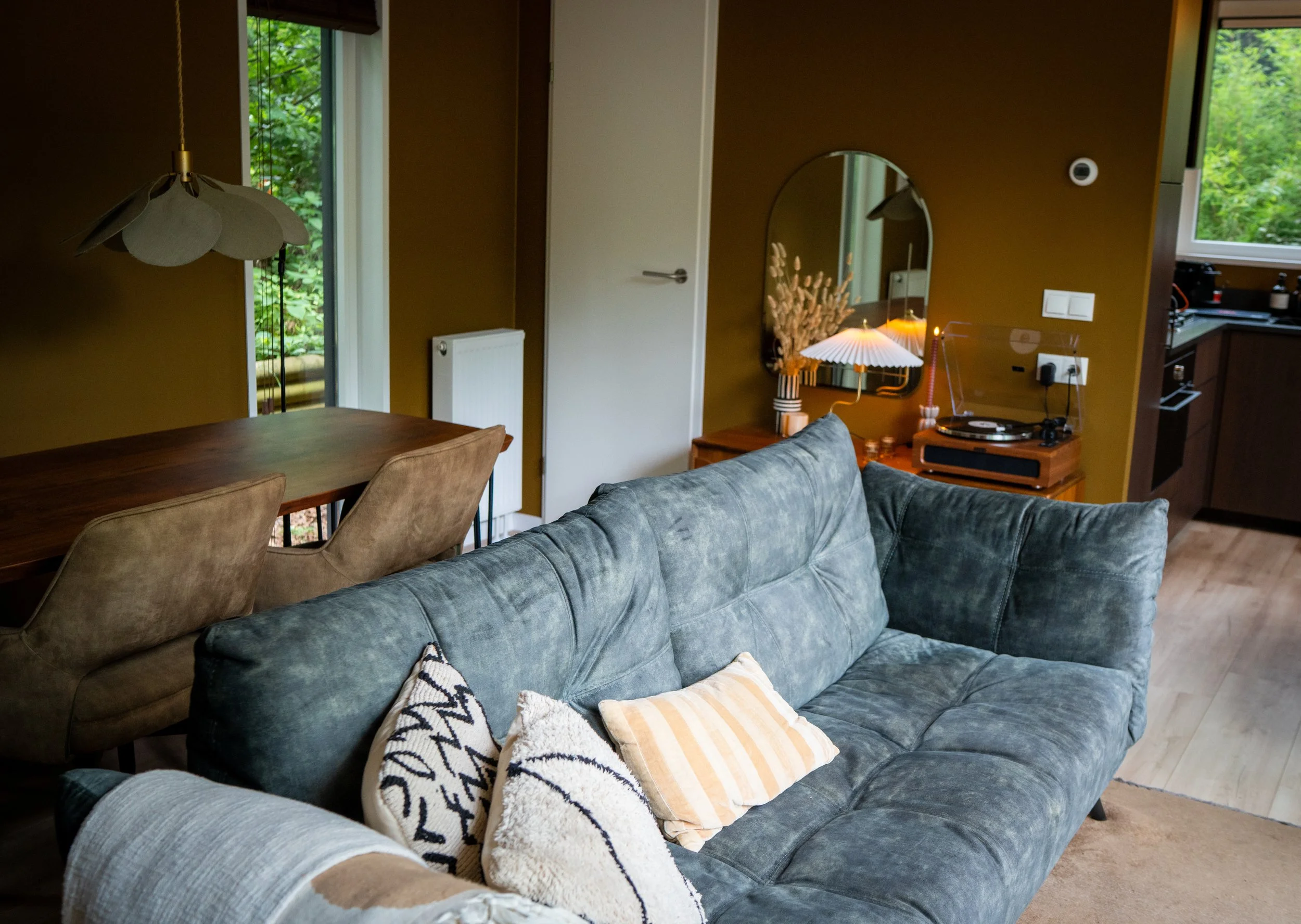 Een woonkamer met een groene bank, decoratieve kussens, een houten tafel, een hanglamp, een spiegel met een vaas met gedroogde bloemen, en een platenspeler, met ramen die naar groene buitentuin uitkijken.