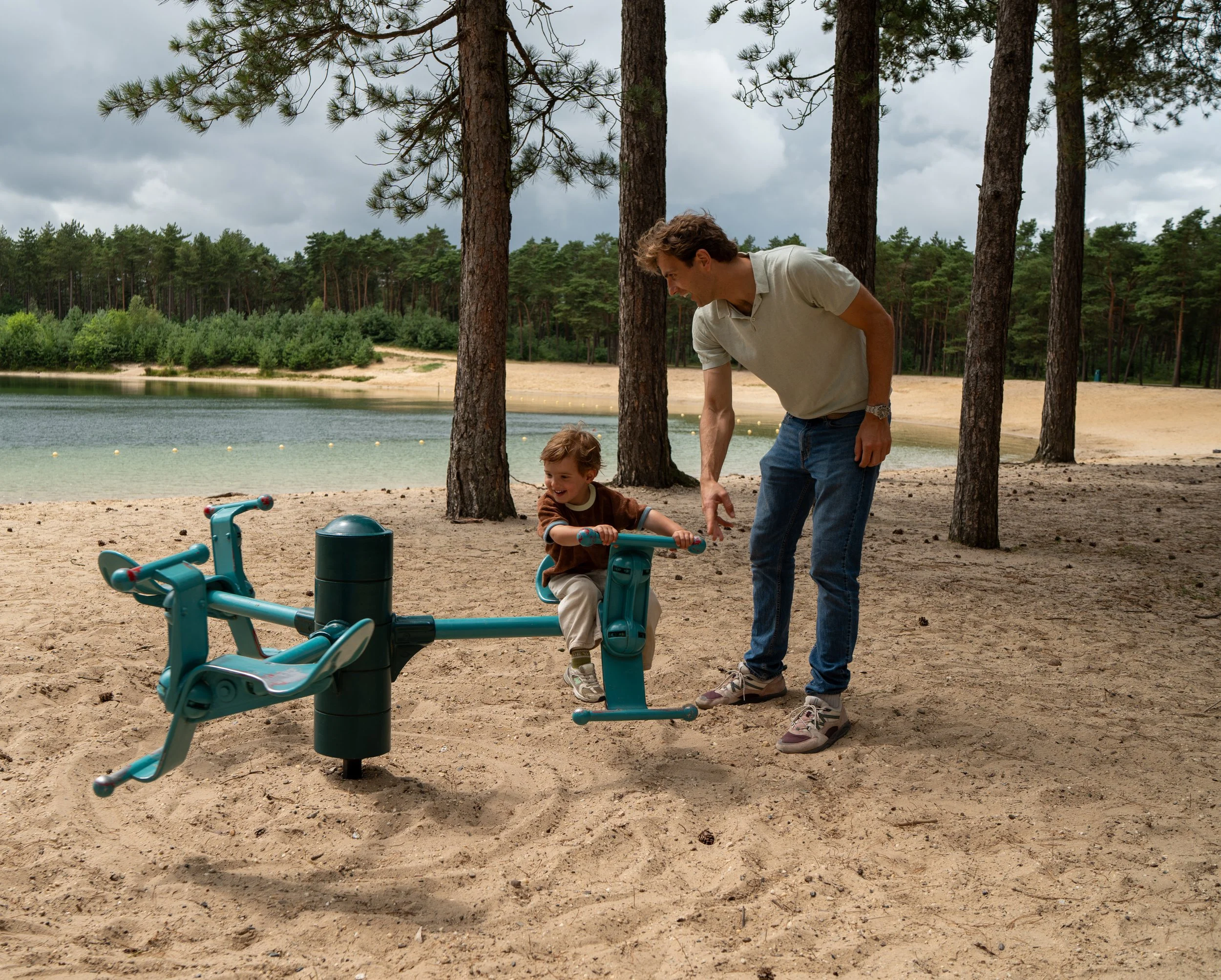 Man en kind spelen bij een waterpomp op een strand, omringd door bomen en een meer.
