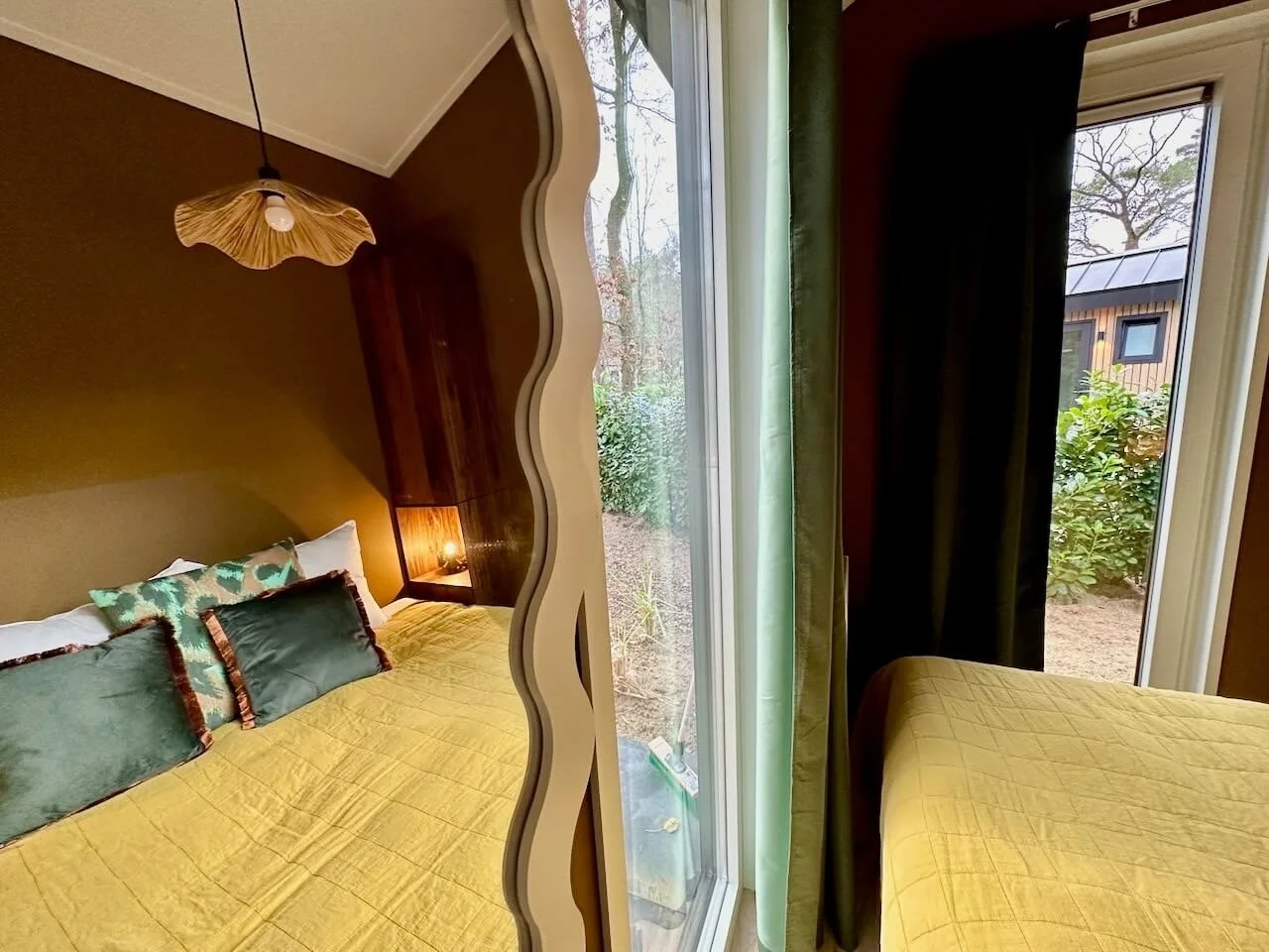 De afbeelding toont een interieur van een slaapkamer met een bed dat bedekt is met een gele deken en decoratieve kussens. Aan de linkerkant is er een spiegel met een golvende rand die het bed weerspiegelt. In de achtergrond is een lamp die uitgaat en een wand met een houten paneel. Aan de rechterkant is een groot raam dat uitkomt op een tuin met struiken en een ander gebouw in de verte.