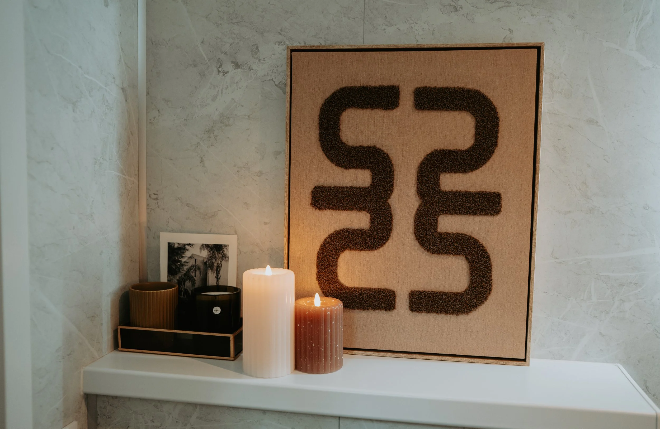 Decoratieve muurkunst met zwart patroon op beige achtergrond, naast kaarsen en plantenbak op witte muur.