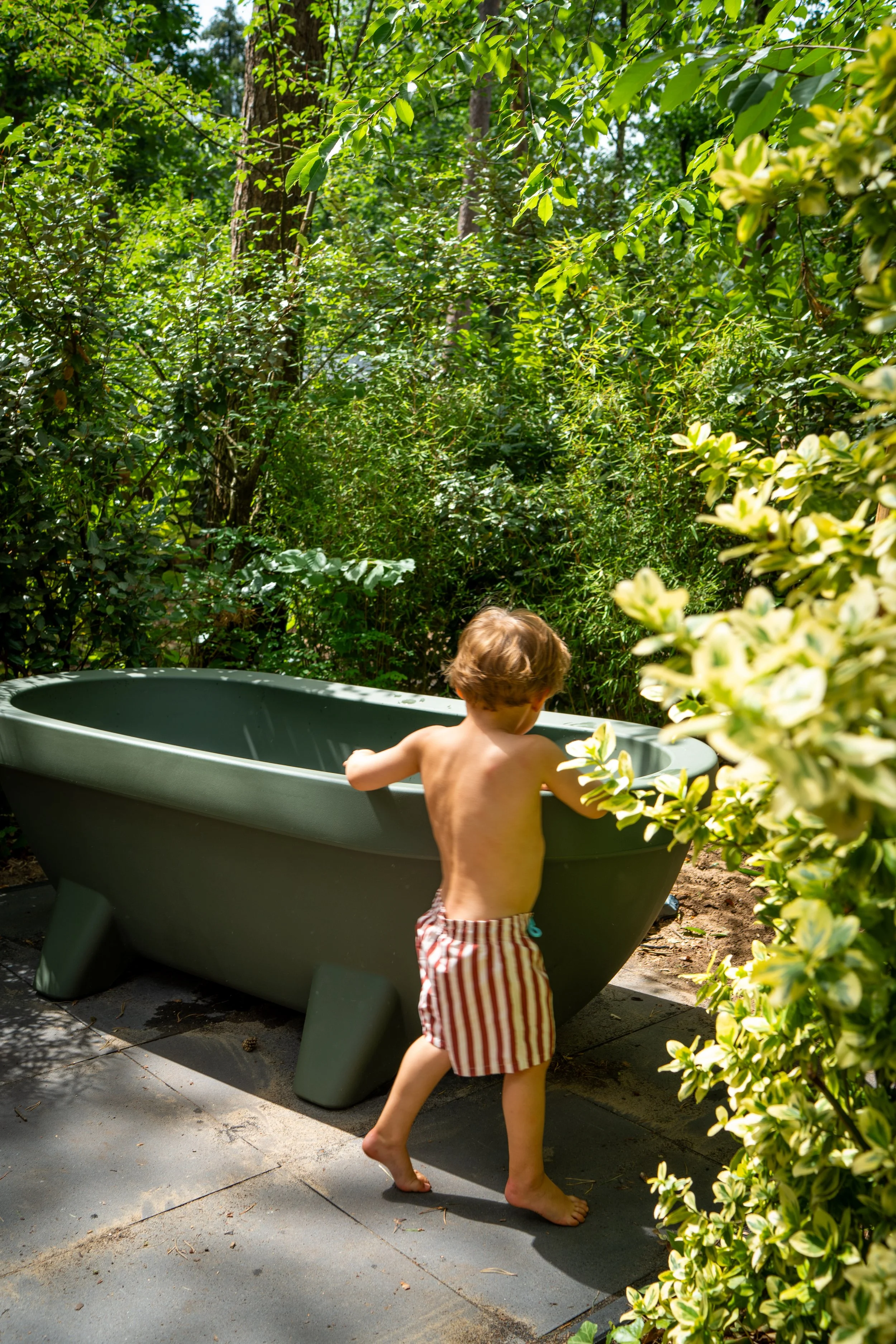 Een kind met zwemkleding die uit een groene wastrog in een tuin met veel groen staat.