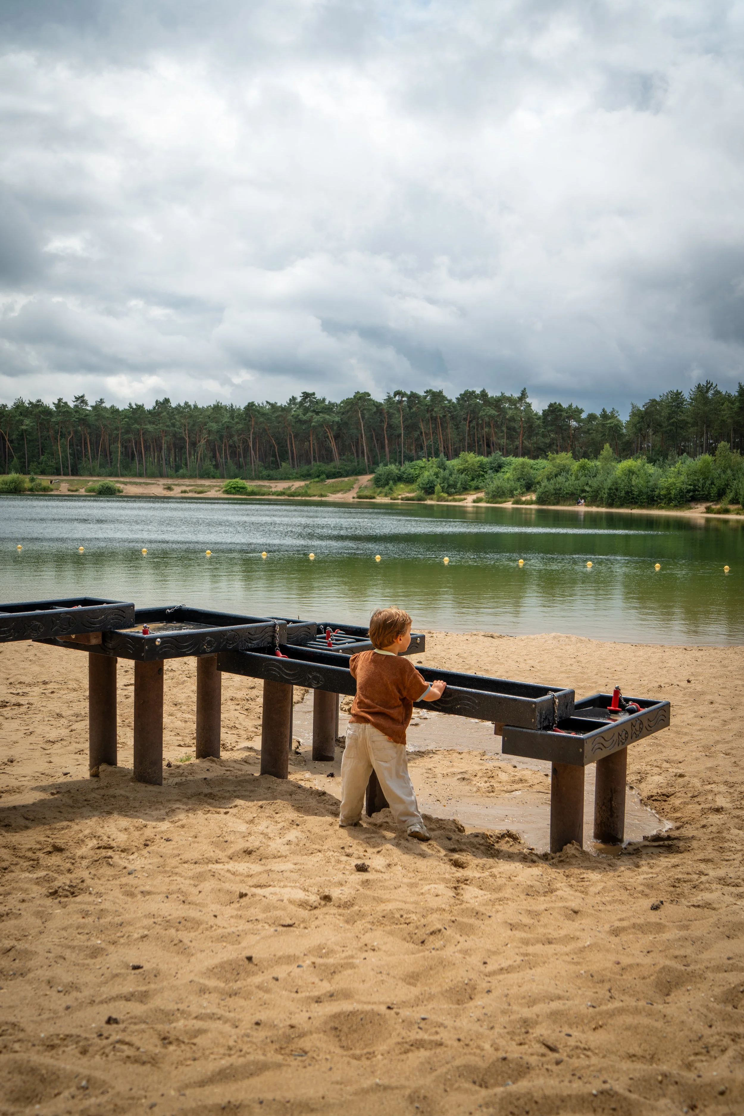 Een kind speelt op een zandstrand bij een meer, met een rij boten in de voorgrond en een bos met donkere wolken op de achtergrond.