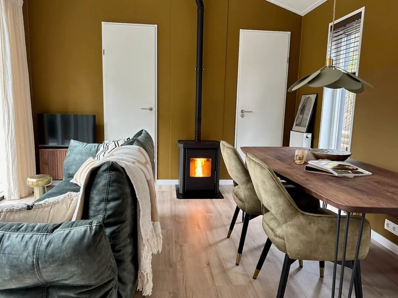 Woonkamer met bank, eetkamertafel, stoelen, houtkachel, tv en raam.