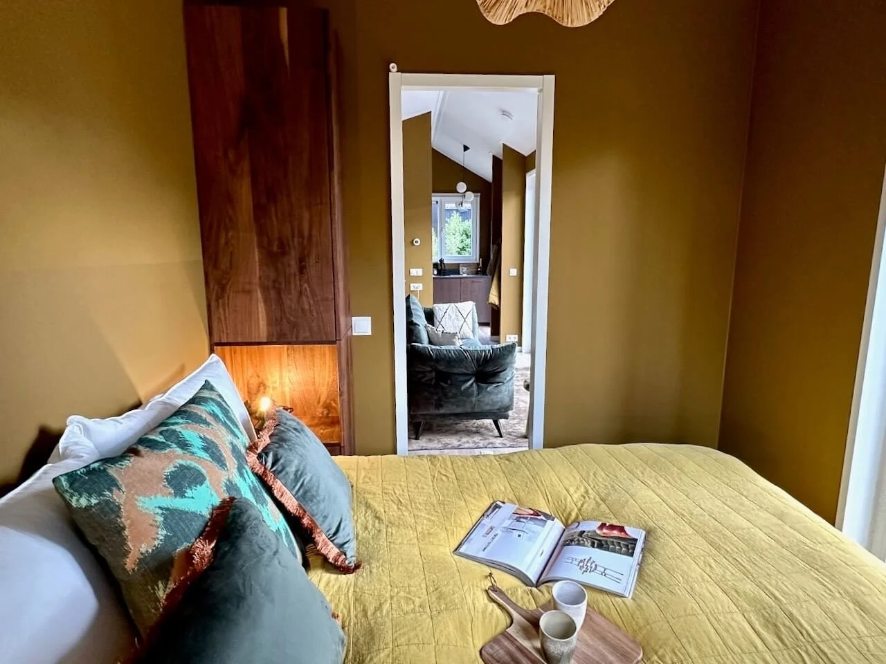 Een slaapkamer met een geelbed, enkele decoratieve kussens, een open boek, en twee bekers op een dienblad. In de deuropening is een zithoek met een donkere fauteuil en een raam dat uitkijkt op groen landschap.