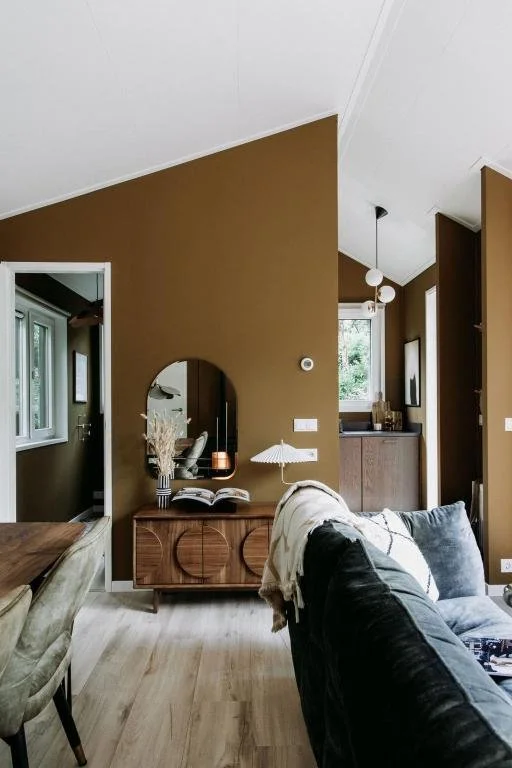 Interieur van een woonkamer met een bruine muur, een grote spiegel, een deco-tak, een open boek op een houten kast, een lamp met een parasolvorm, een bank met kussens en een aanvankelijke keuken met hanglampen in de achtergrond.