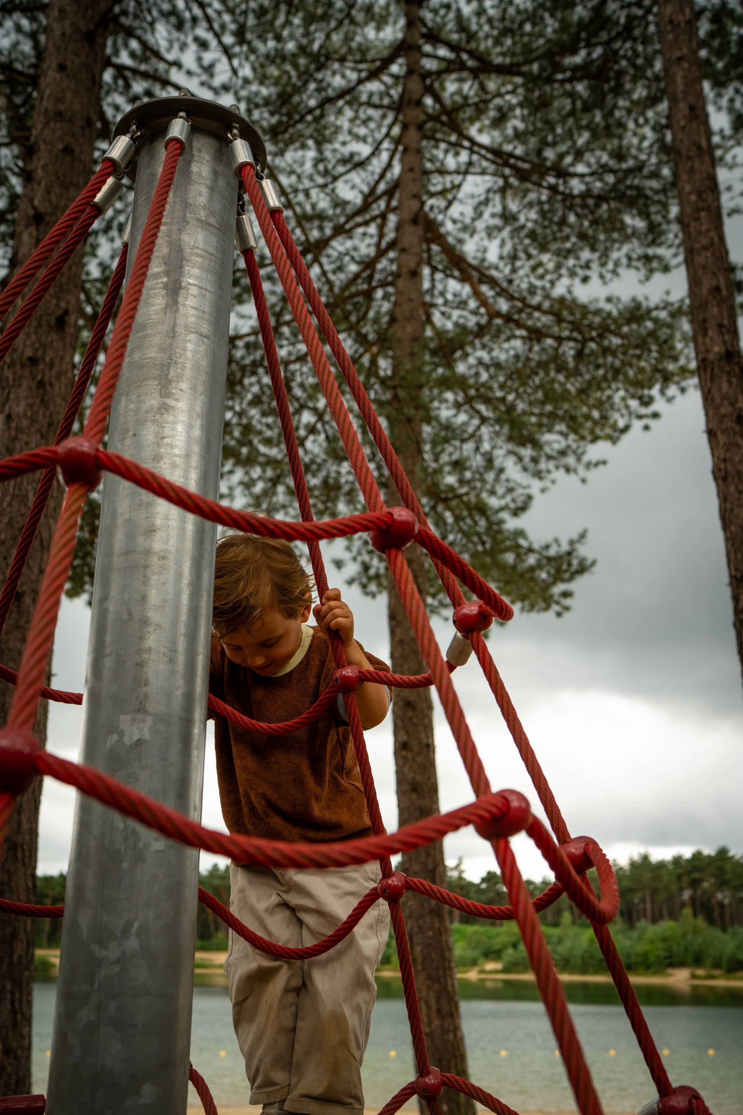 Een kind dat op een rope klimnet speelt in een bosrijke omgeving met water en bomen op de achtergrond.