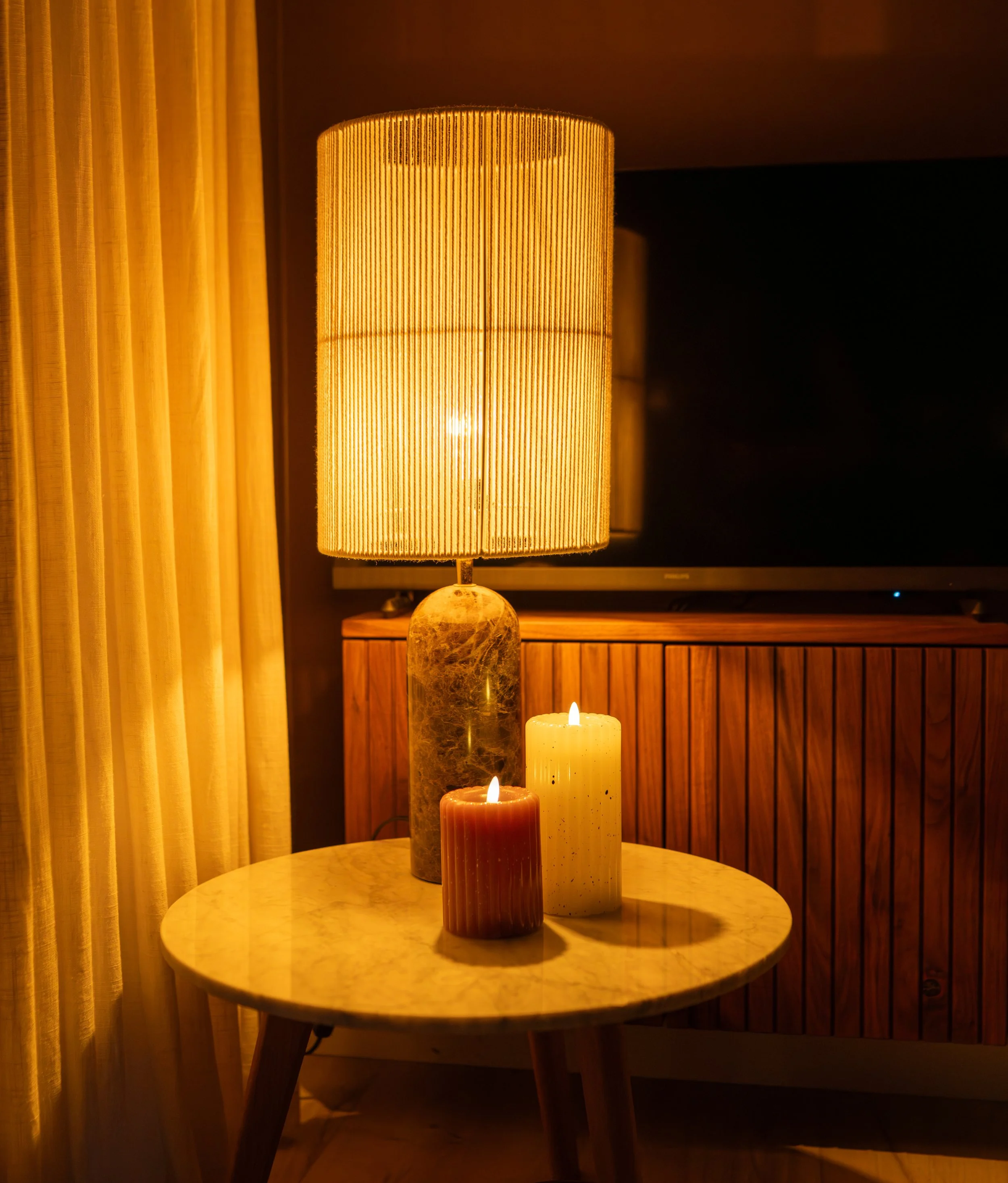 Een ronde tafel met vier brandende kaarsen in verschillende kleuren en maten. Op de tafel staat een lamp met een oranje gecoate kappen, daarnaast staan gele, witte en rustkleurige kaarsen. In de achtergrond een tv en een houten kast.