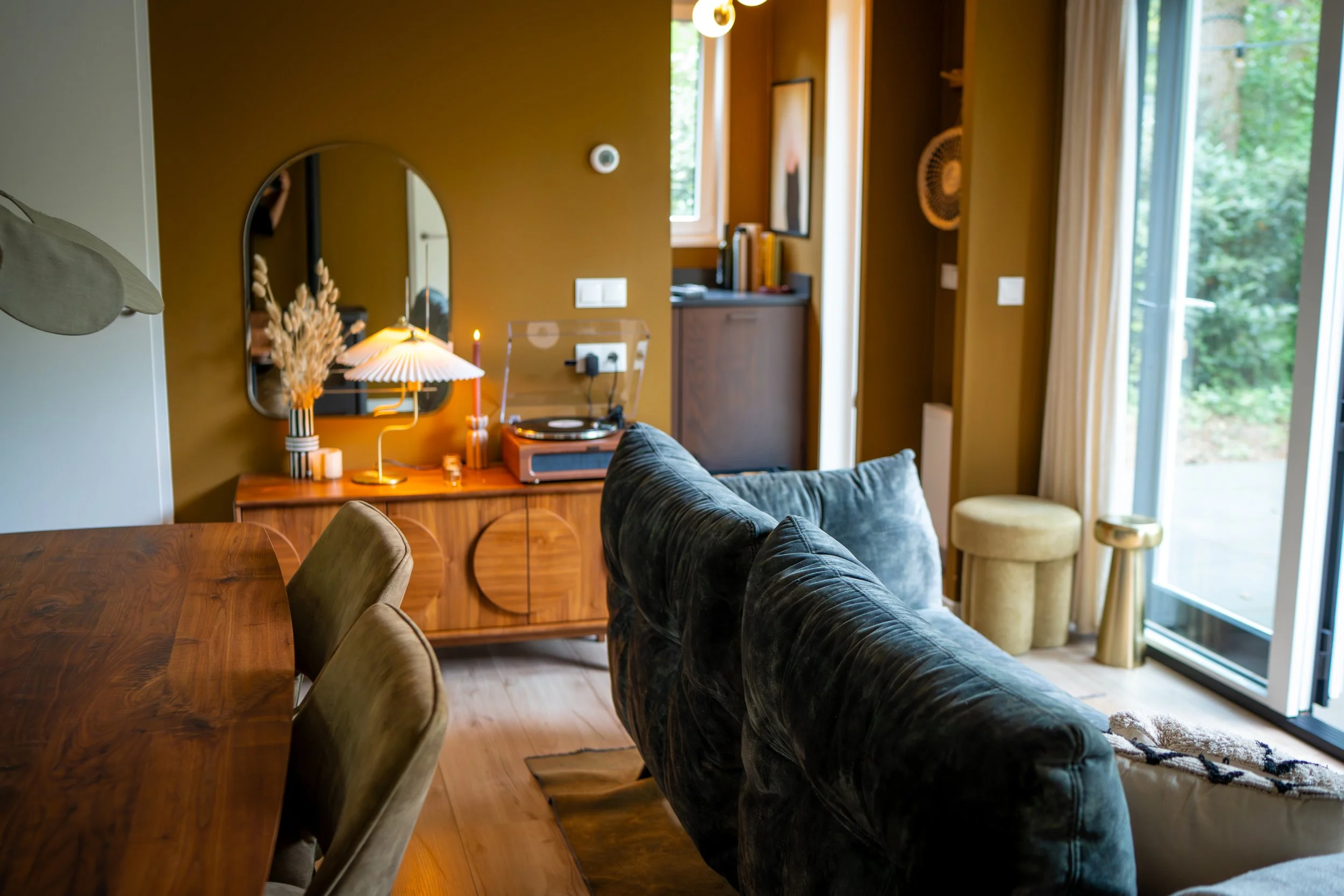 Een gezellige woonkamer met een houten eettafel, zachte zittingen en een vintage platenspeler, met grote ramen die uitzicht bieden op groene bomen buiten.
