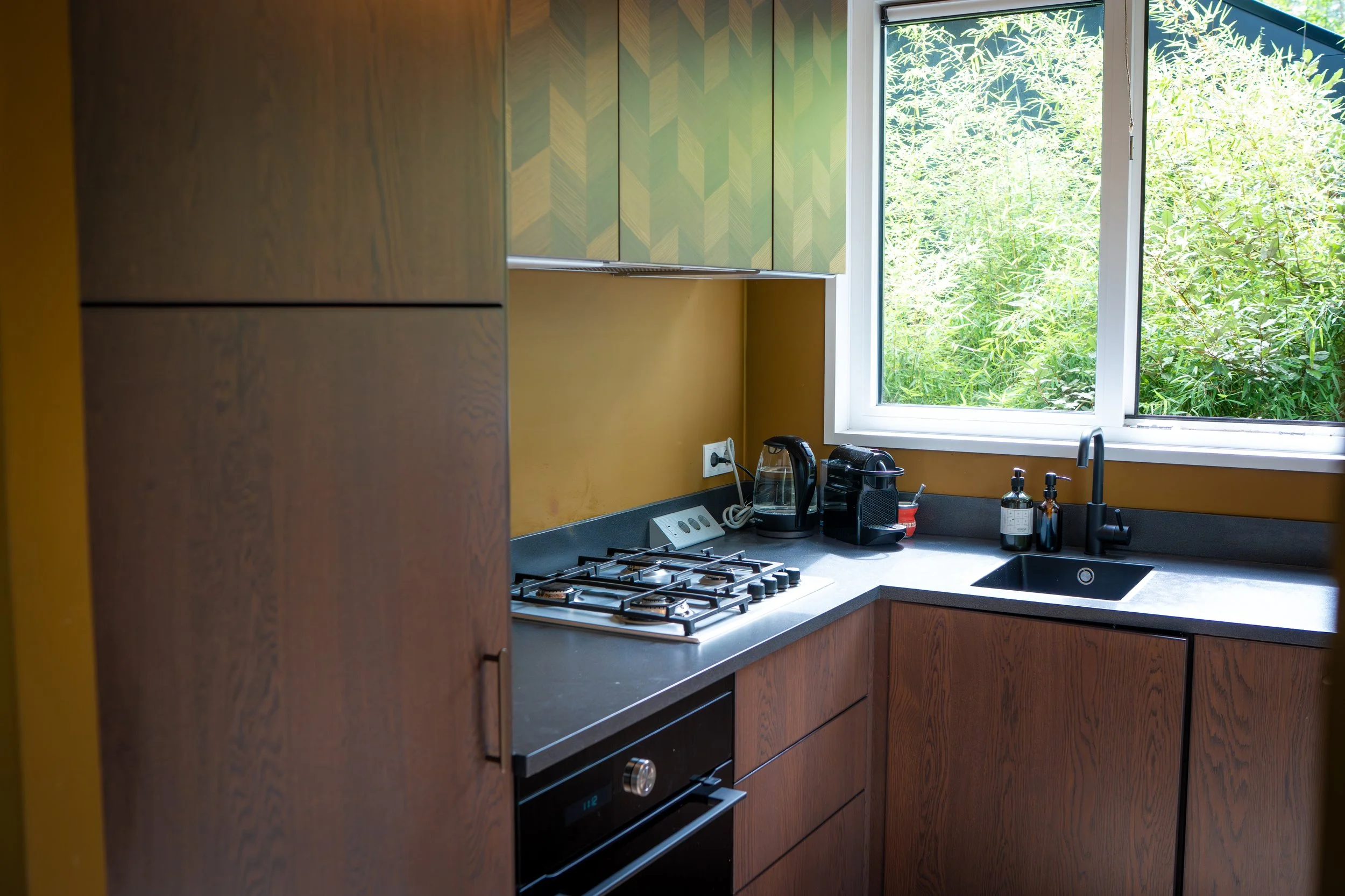 Een keuken met houten kasten, een gasfornuis, een gootsteen, een zwarte kraan en verschillende kleine apparaten en pexample in de gootsteen bij een raam dat uitkijkt op groene planten.