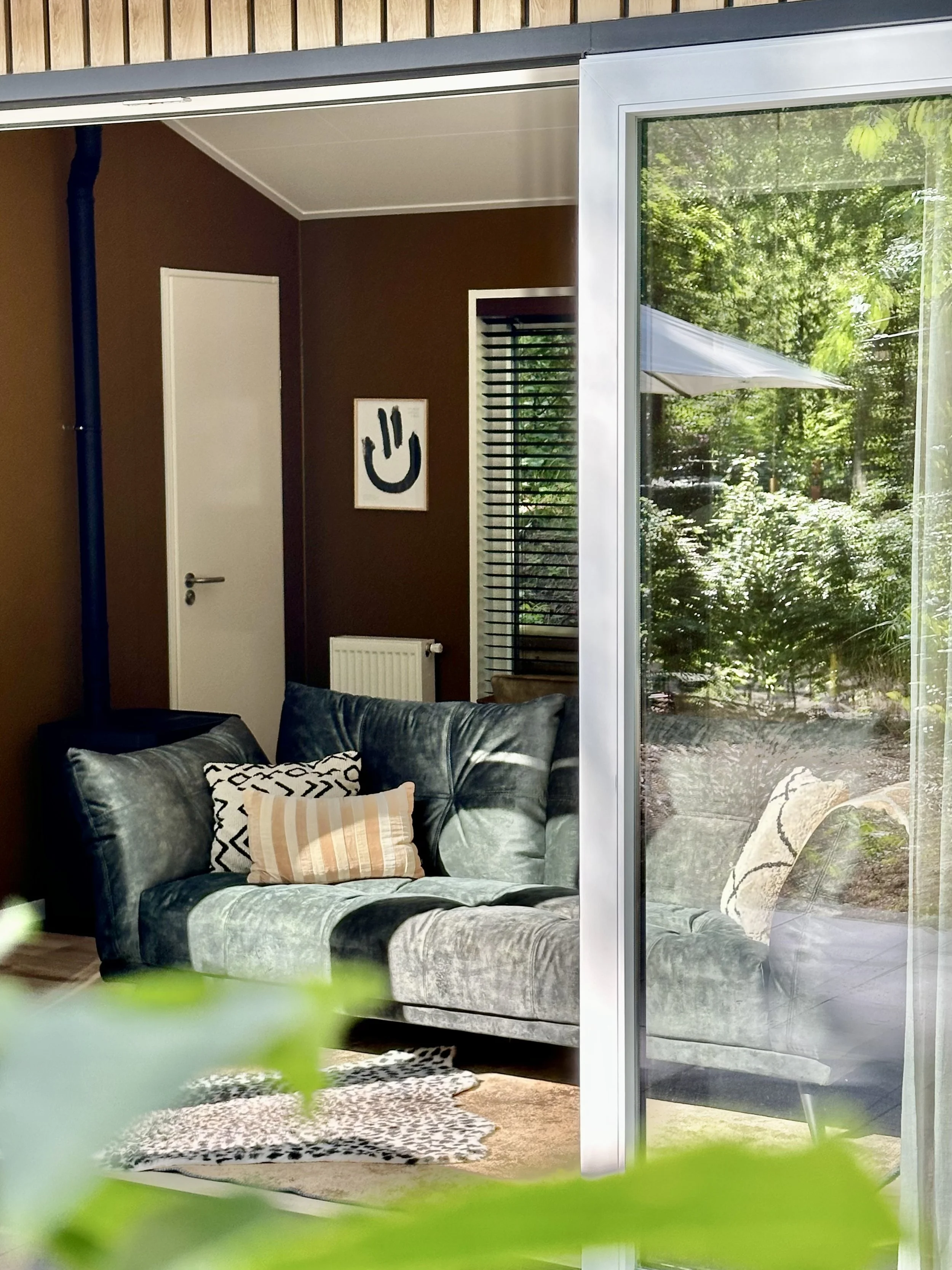 Binnenruimte met een groene bank, meerdere kussens, een muur met een kunstwerk en een grote glazen muur naar een tuin met bomen en een parasol.