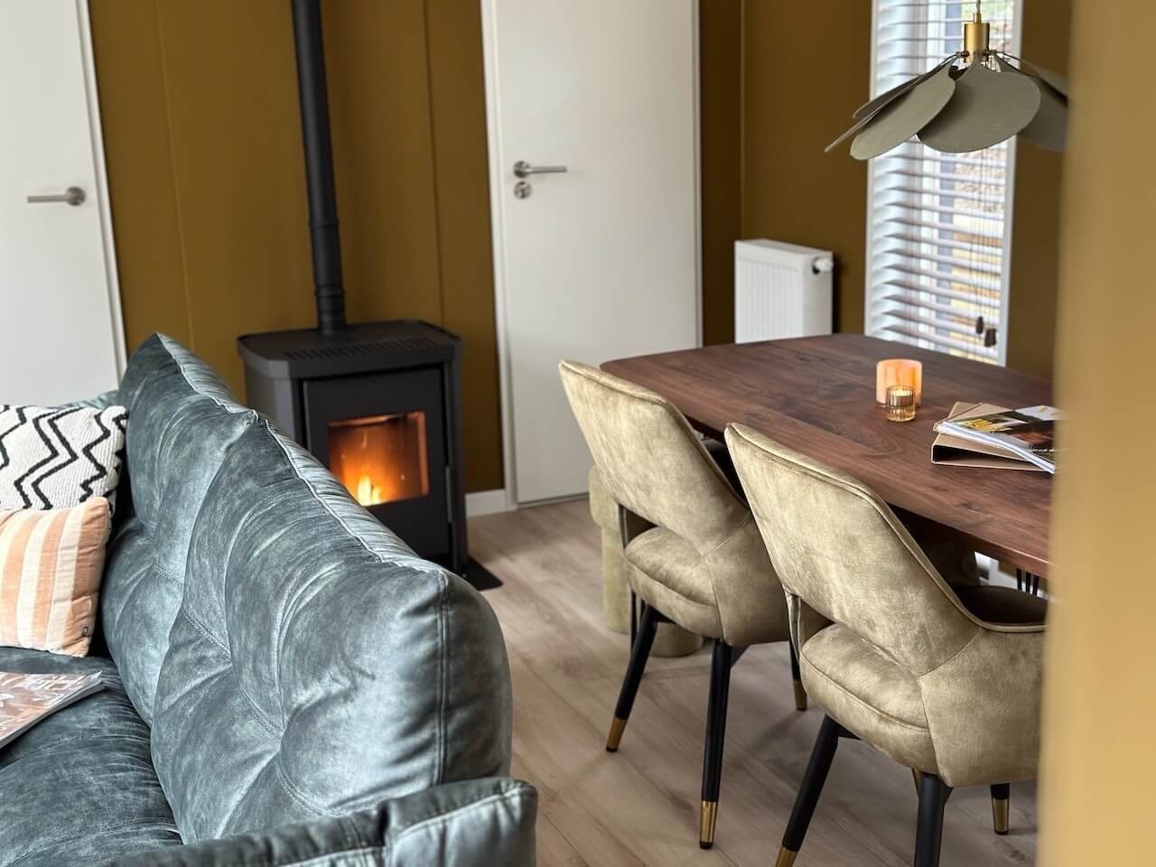 Een woonkamer en eethoek met een bank, een houtkachel die brandt, een houten eettafel met beige stoelen, een lamp met bladeren, kaarsen en tijdschriften op de tafel. Er is een grote raam met jaloezieën.