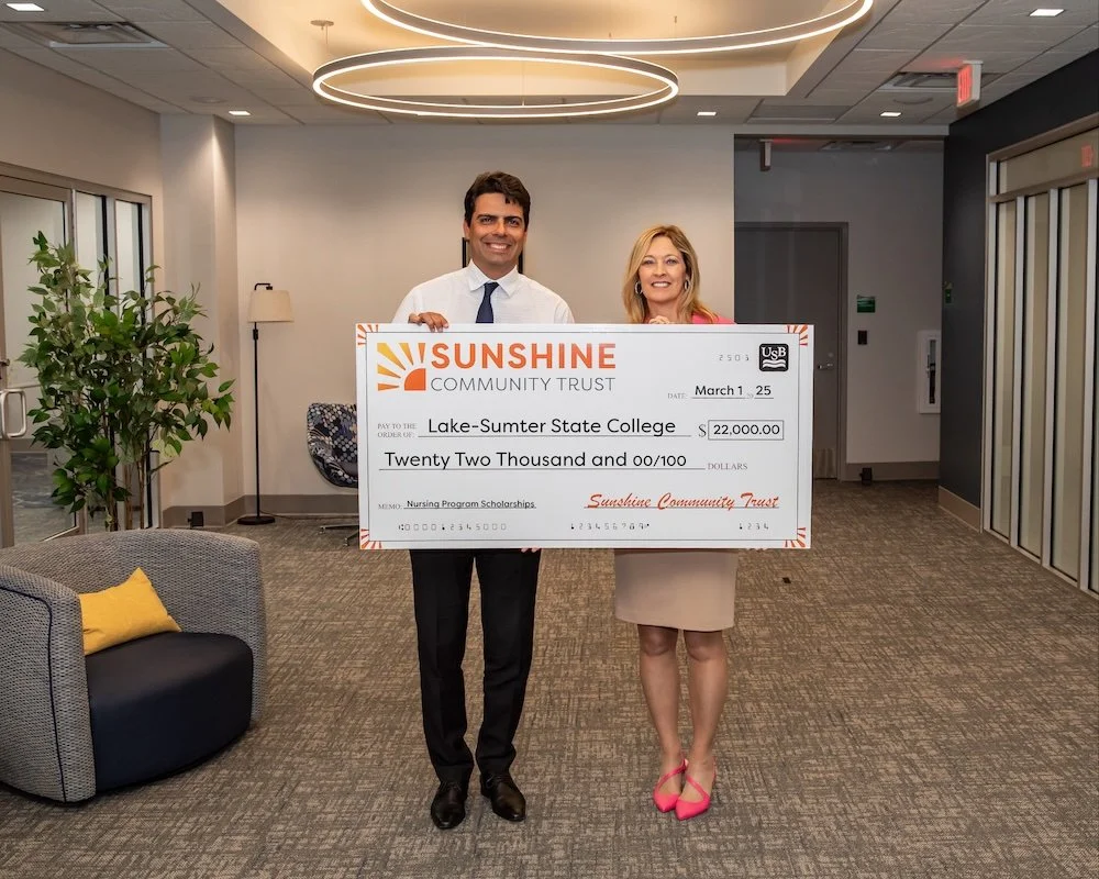 Two people holding a large check from Sunshine Community Trust made out to Lake-Sumter State College for $22,000, standing in a modern office with a plant and a gray chair.
