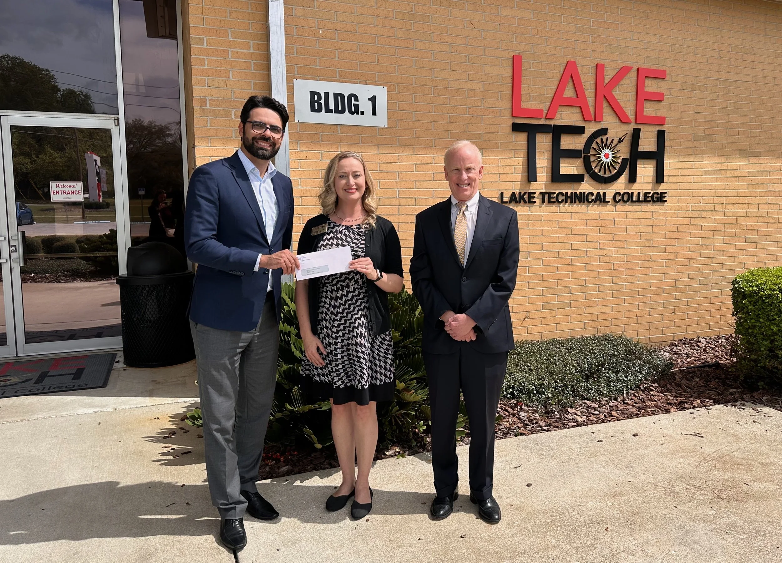 Three people stand outside a building labeled Lake Technical College, with a sign indicating Building 1. The group includes a man in a blue suit, a woman holding a check, and an older man in a dark suit. They are smiling and dressed in professional attire.