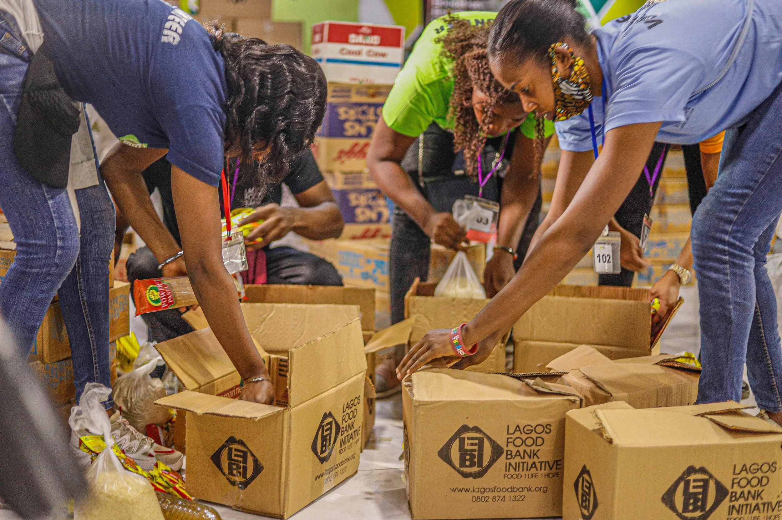 People packing food into boxes at a warehouse, with stacks of boxes in the background.