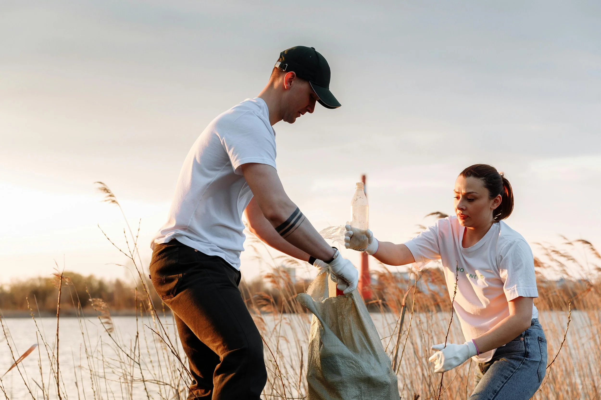 Two people working together outdoors during sunset, cleaning up in a field with tall grass, one man wearing a black cap and white t-shirt, a woman wearing a white T-shirt and jeans, both handling a large net or bag.