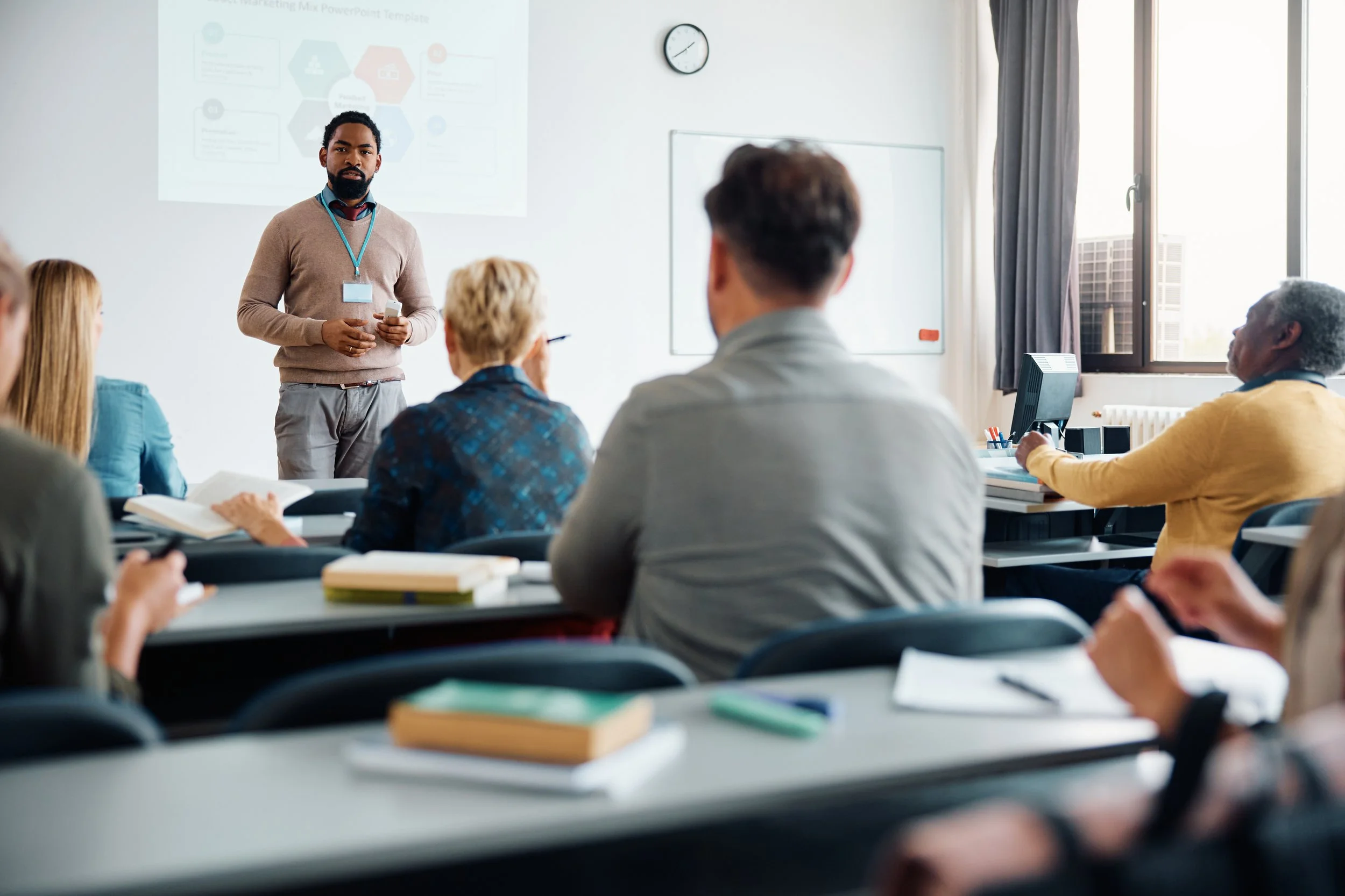 adult students in a lecture hall listen to an African American professor at the front of the room. He is wearing a khaki sweater and a name badge and standing in front of a digital presentation.