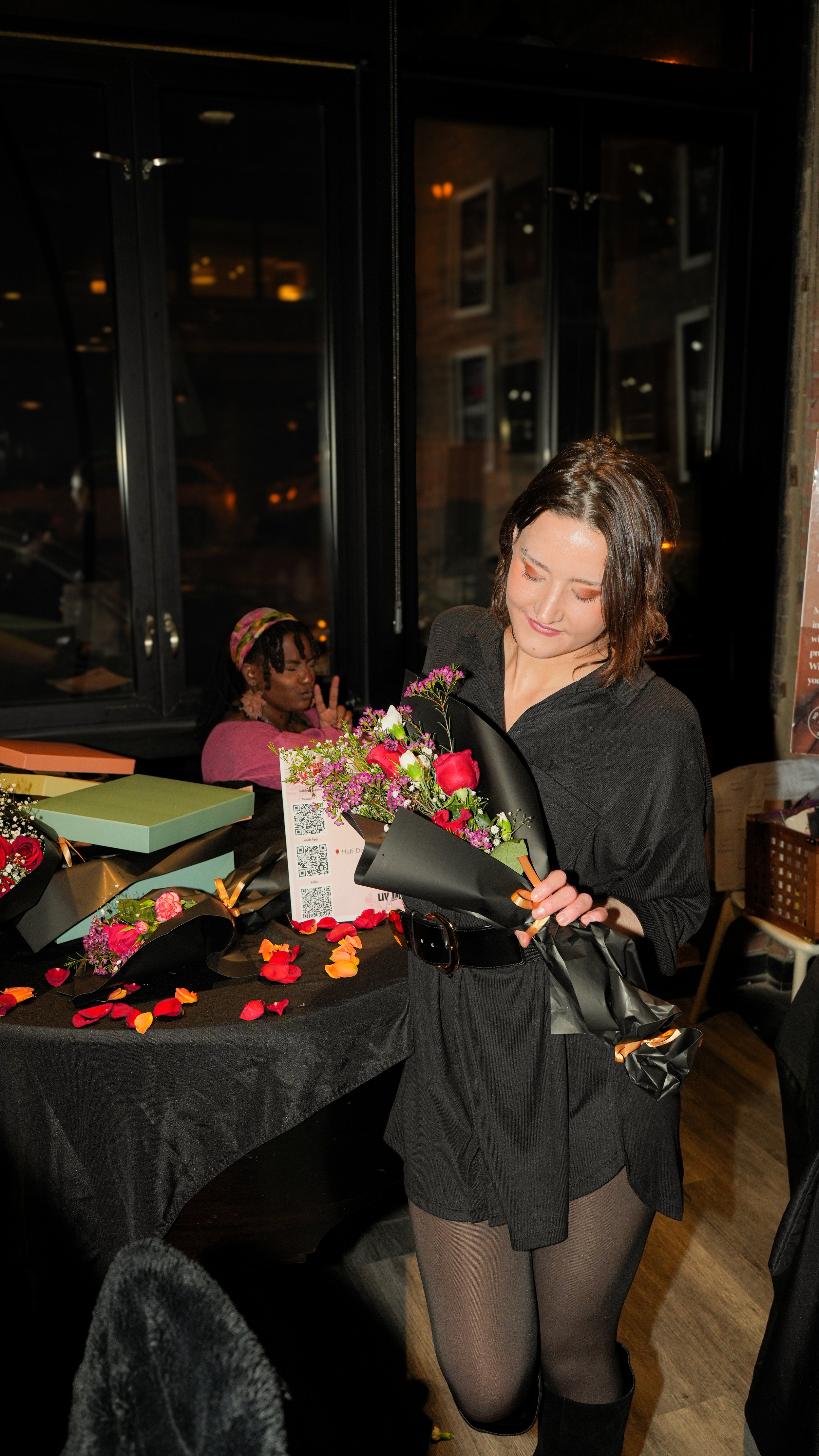 Woman holding a bouquet of flowers at a celebration event, with a table of flower arrangements and rose petals in front of her, in a dimly lit room.