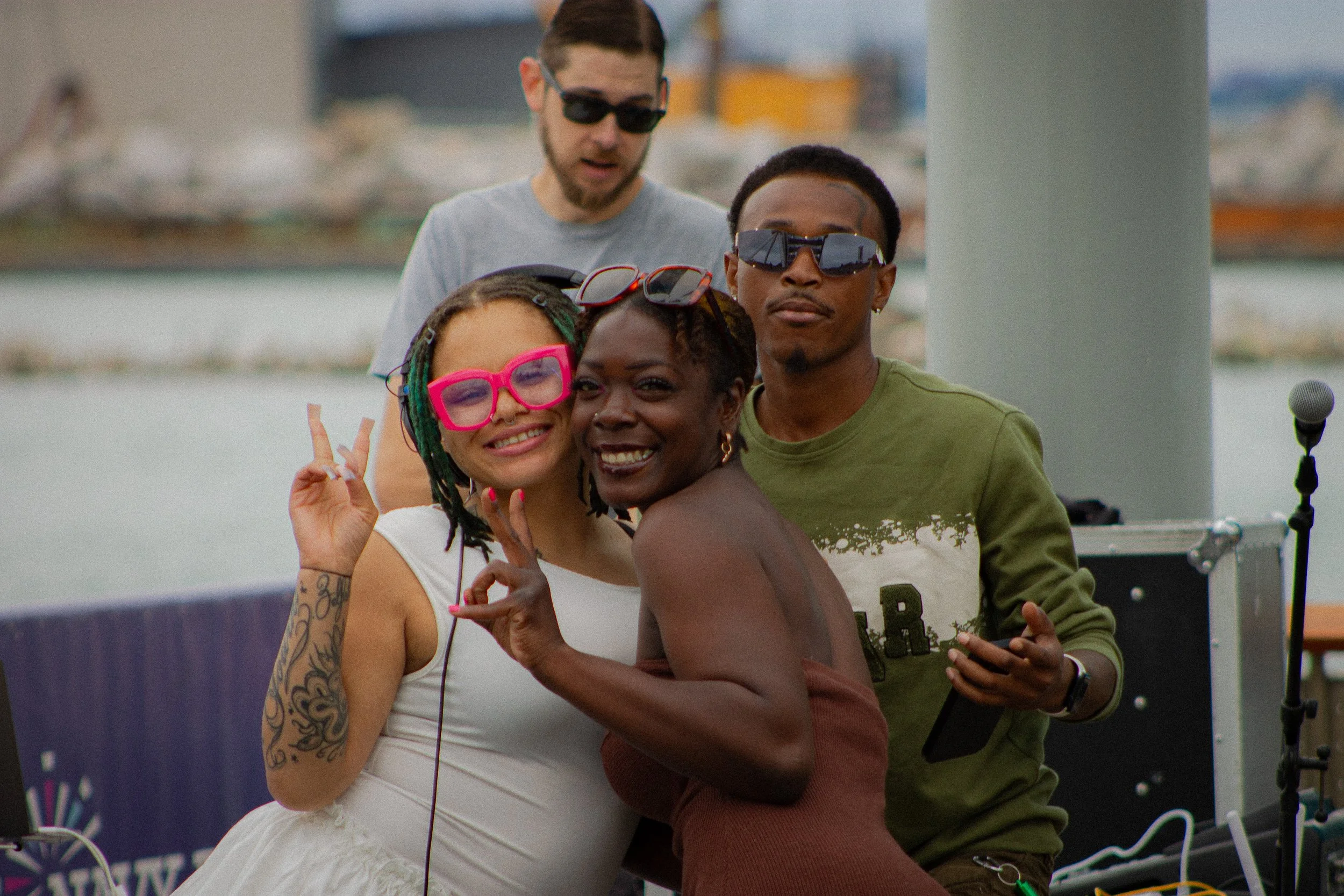 Four people posing together outdoors, three of them wearing sunglasses, smiling, and making peace signs, with a microphone in the foreground.