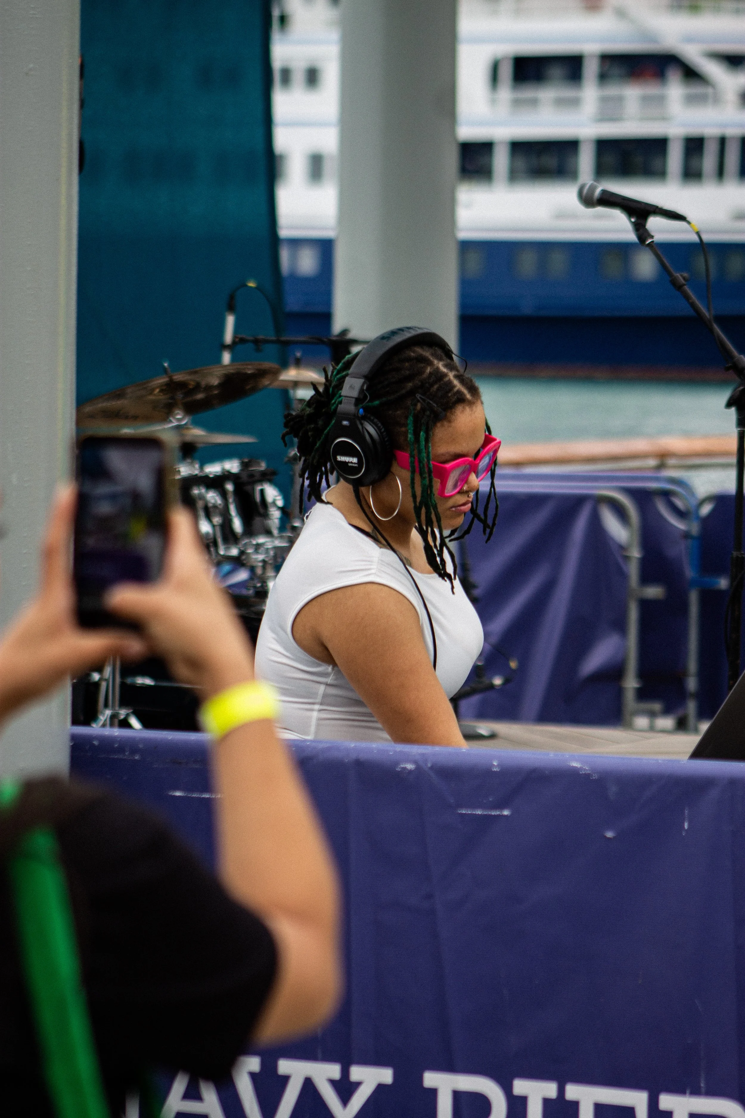A woman with dreadlocks wearing pink sunglasses and headphones, standing behind a blue barrier with white text, in an outdoor setting near a body of water and a boat in the background.