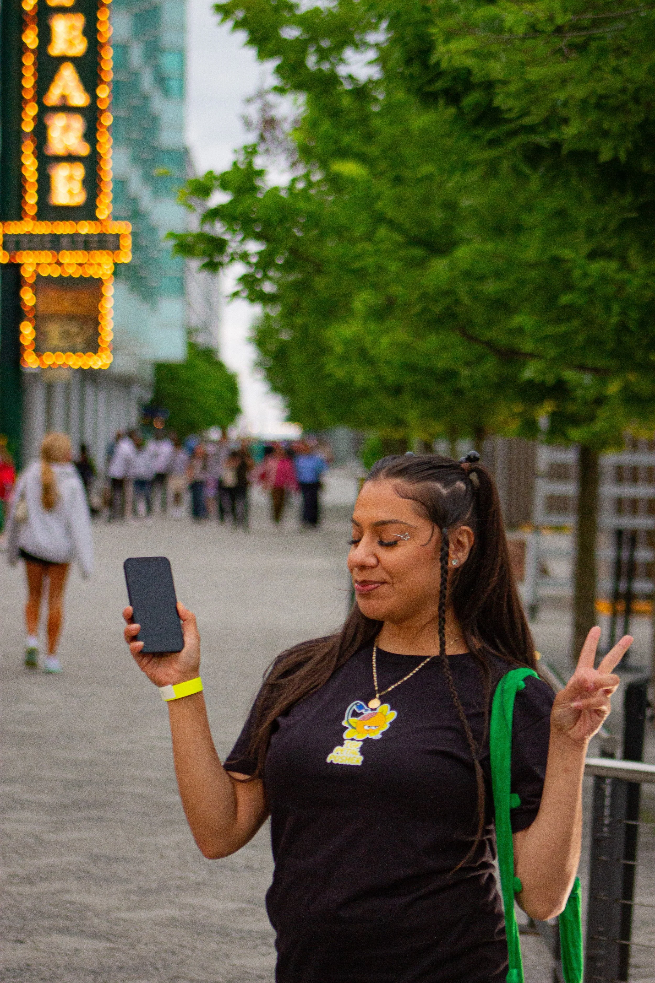 A woman with glasses and braided hair smiling and making a peace sign while holding a smartphone in an outdoor urban setting with green trees and neon signs in the background.