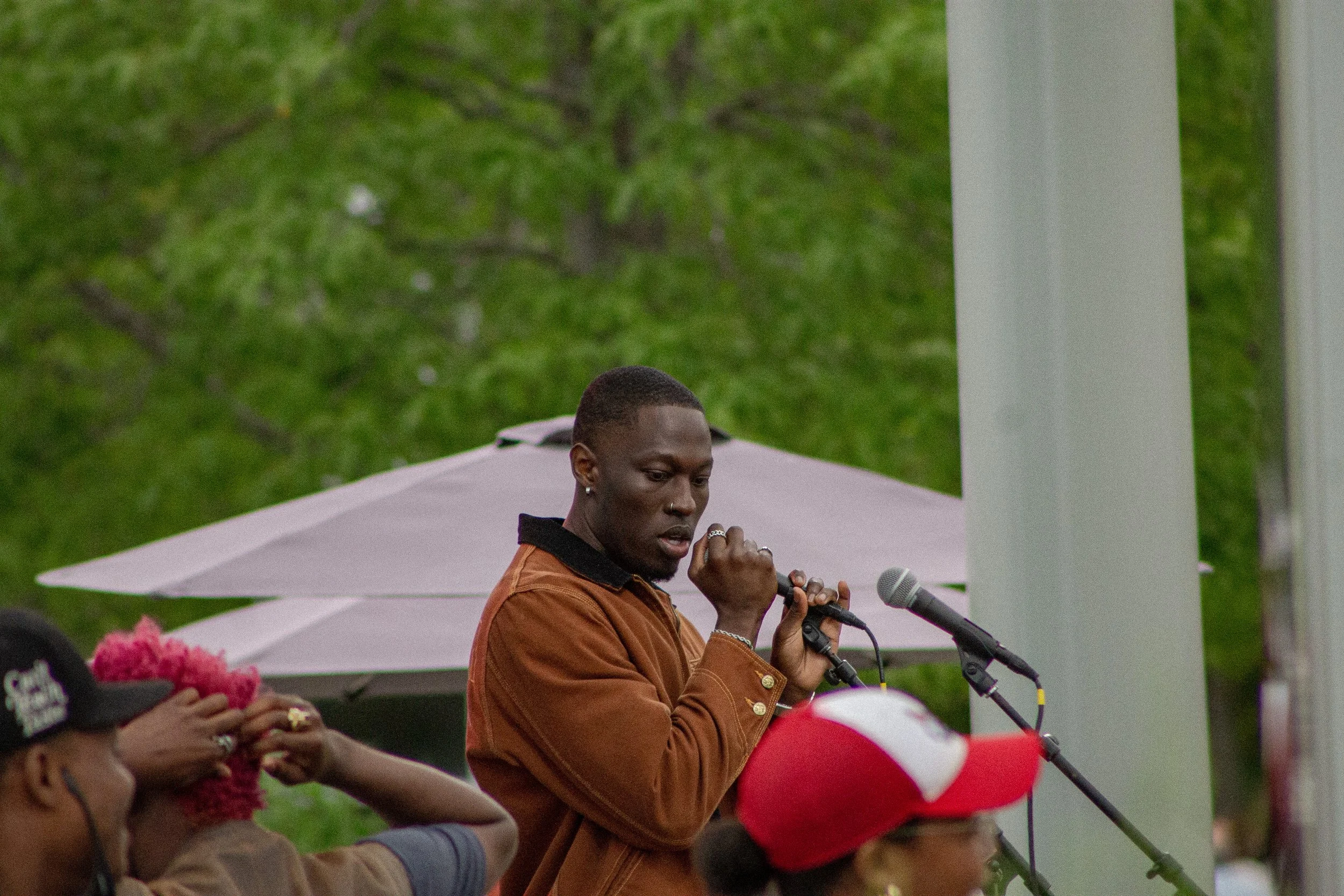 A woman with short hair speaking into a microphone at an outdoor event, with green trees in the background and people holding pink and red balloons.