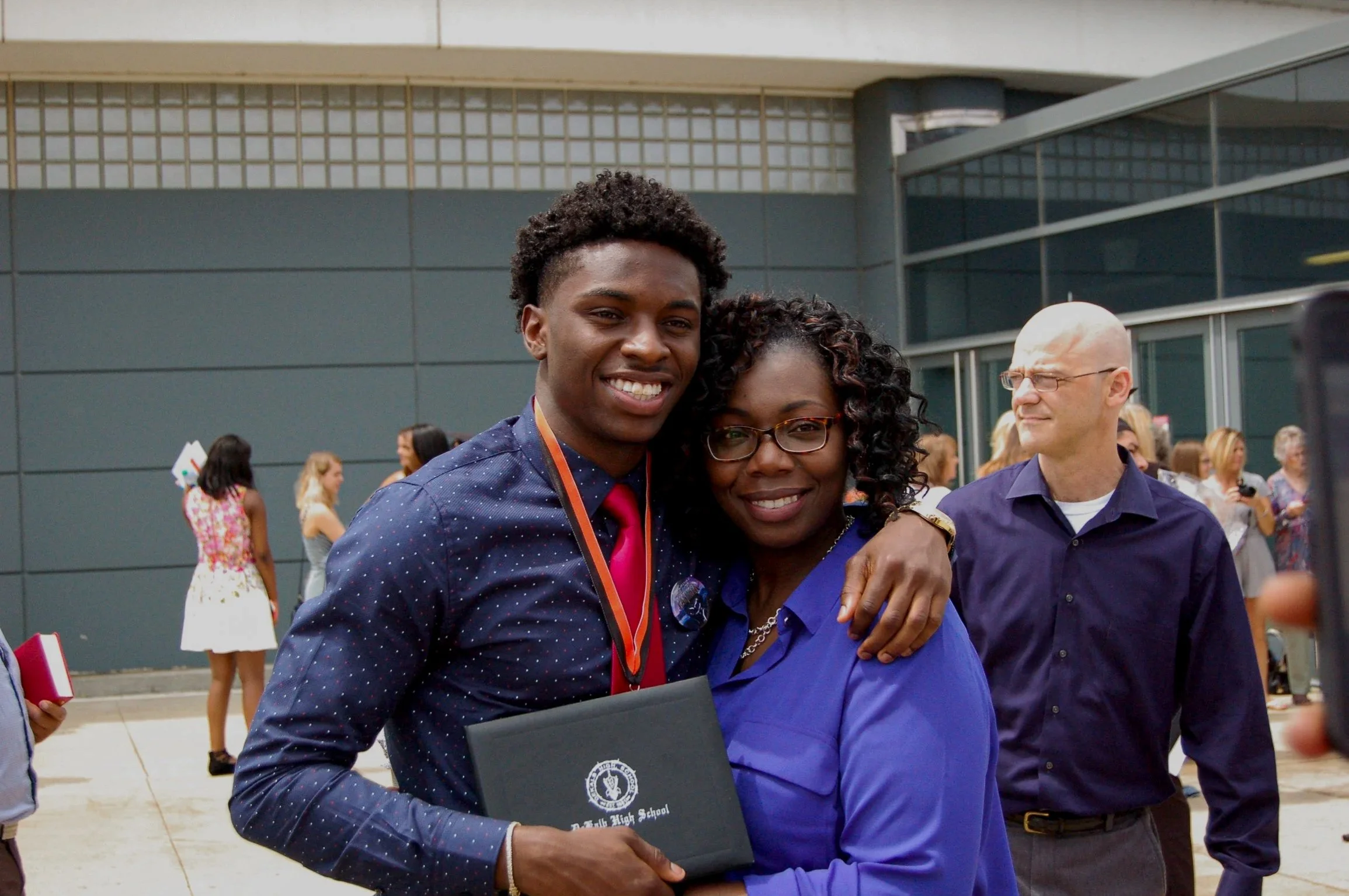 A young man and a woman are embracing and smiling at an outdoor event. The young man is holding a diploma or certificate. There are other people in the background, and a modern building behind them.