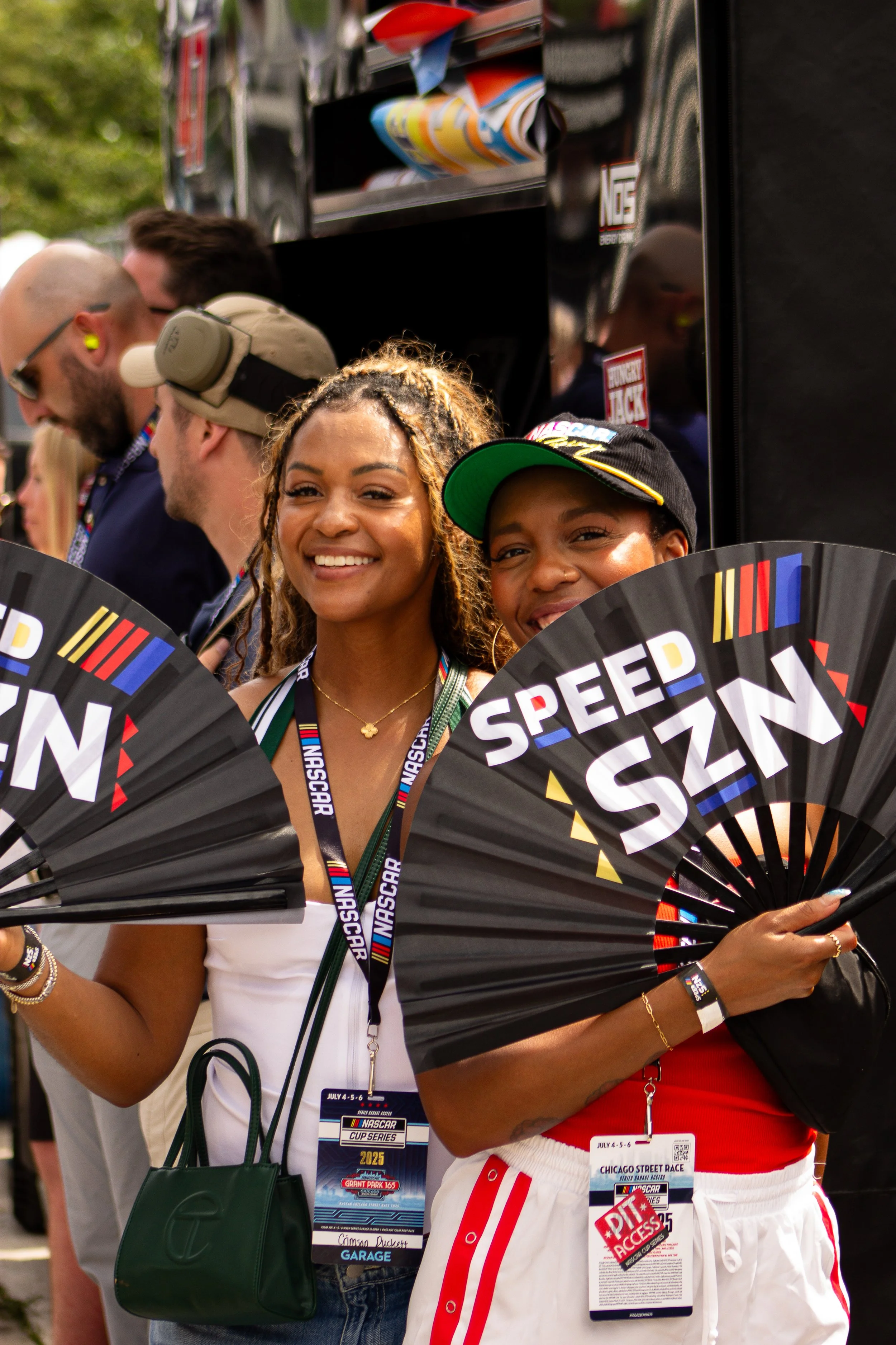 Two women smiling and holding black fans with colorful text, standing at a NASCAR event, with other people in the background.
