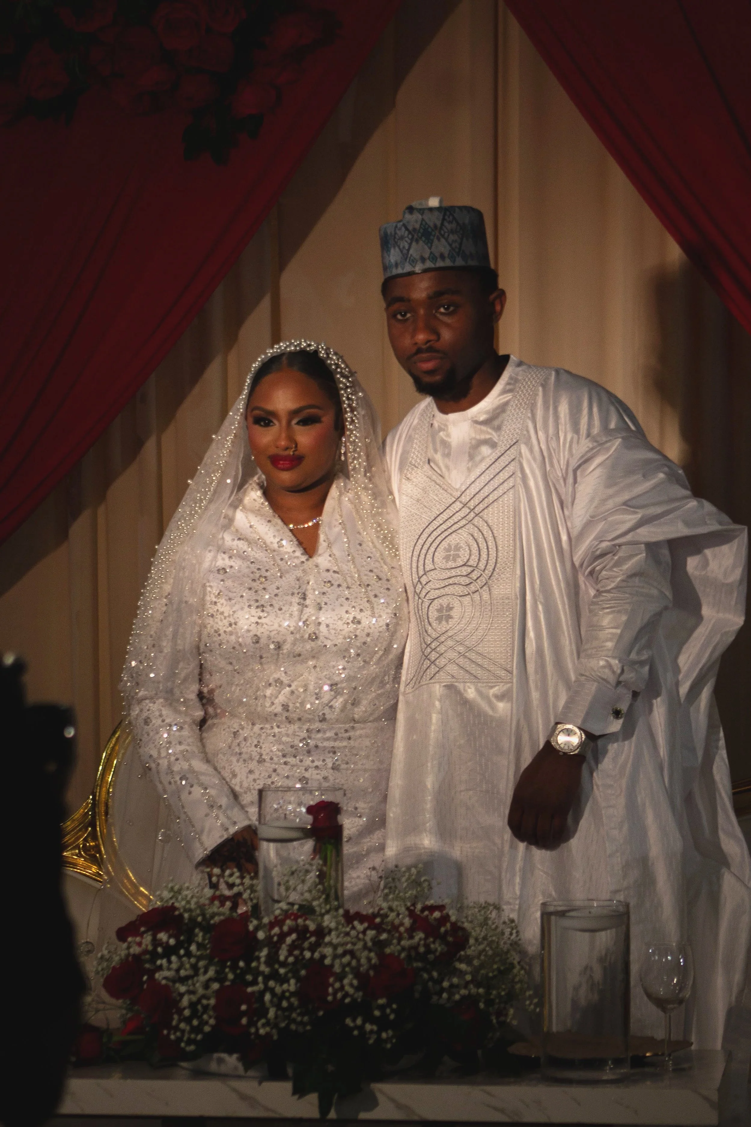 A couple dressed in traditional Nigerian wedding attire, standing together at their wedding ceremony, with floral arrangements and decorations in the background.