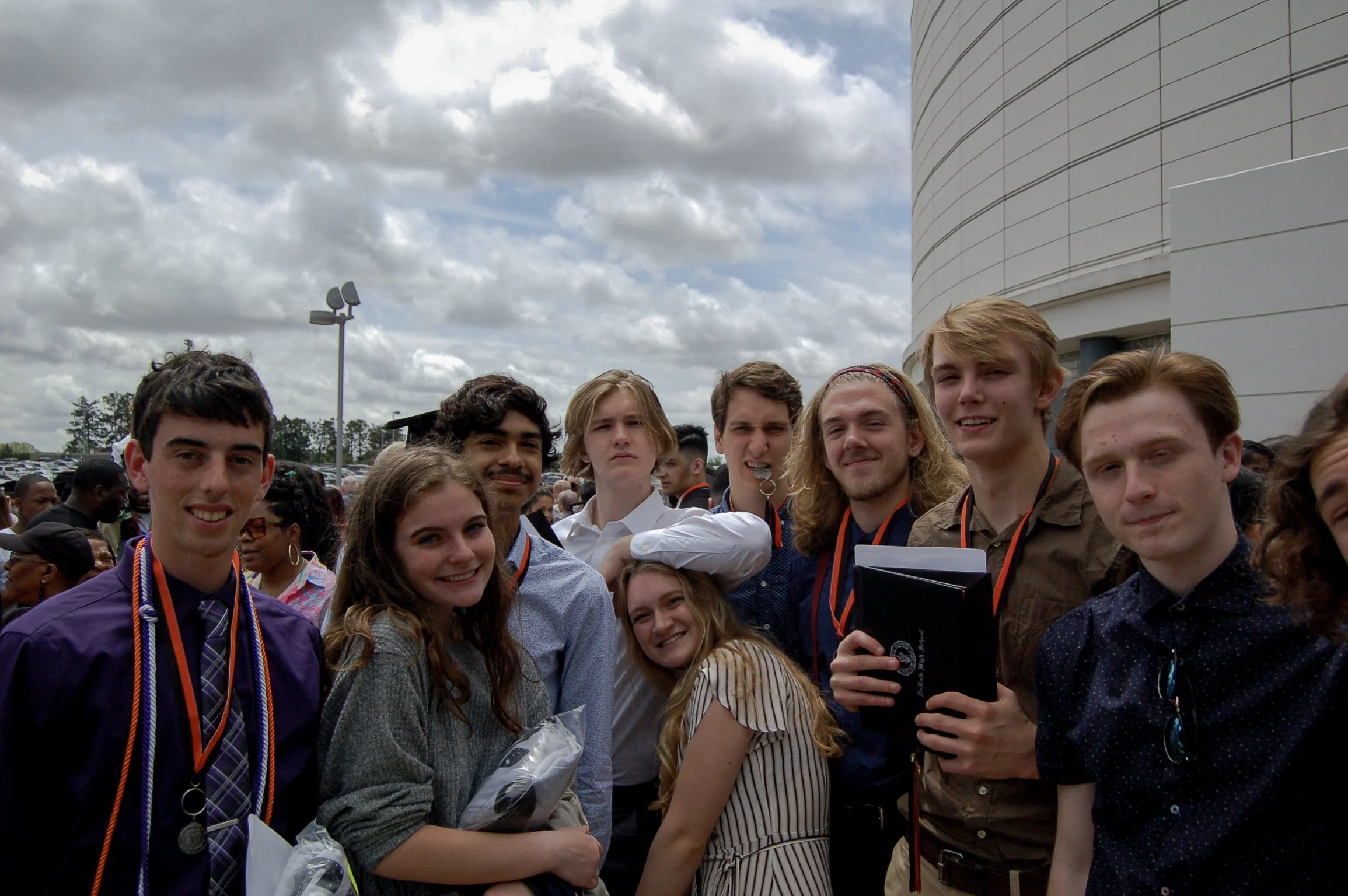 Group of young people gathered outdoors, posing for a photo, with a modern building and cloudy sky in the background.