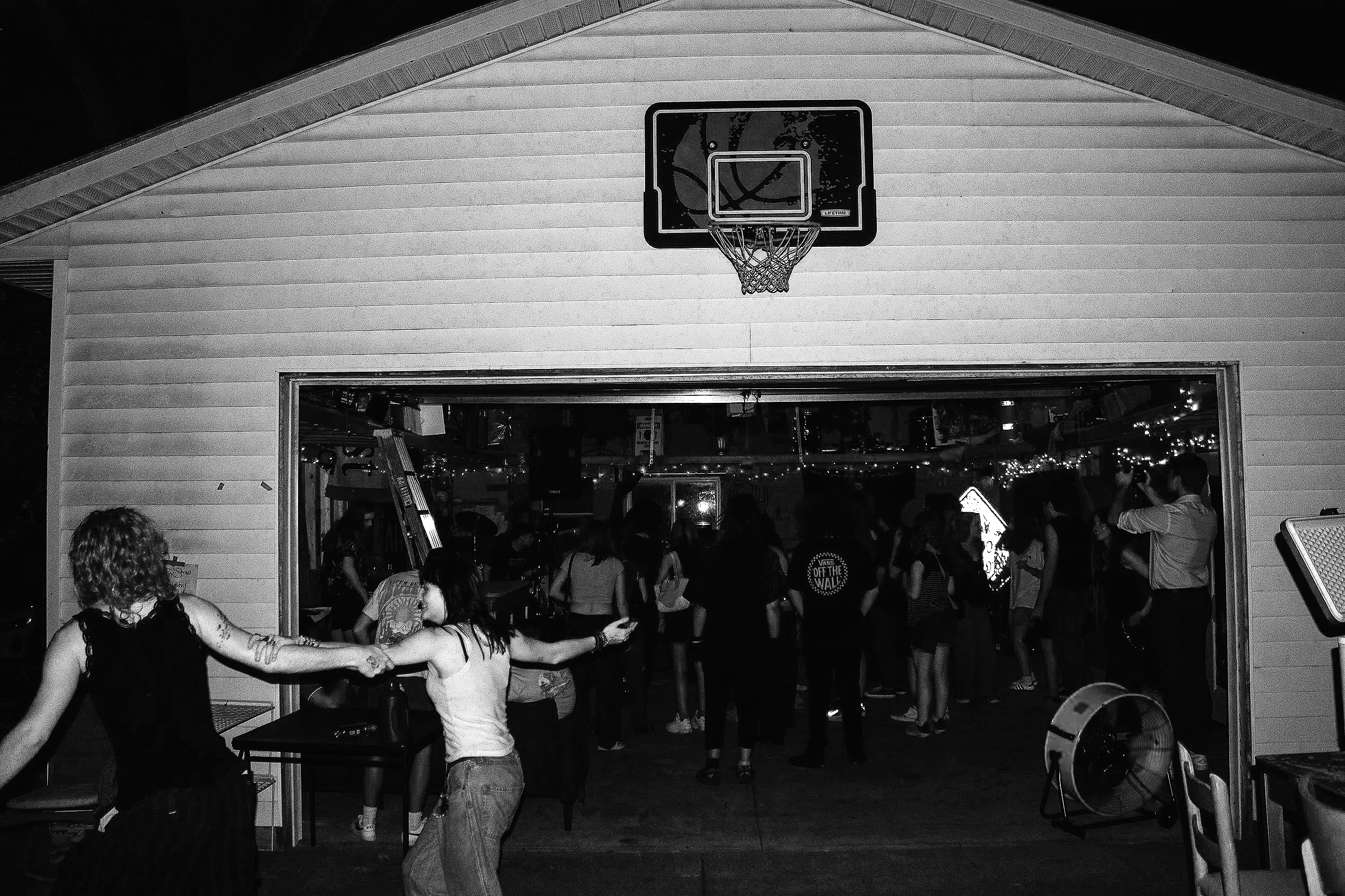 People dancing and socializing indoors at a gathering, with a basketball hoop mounted on the wall above an open space filled with people.