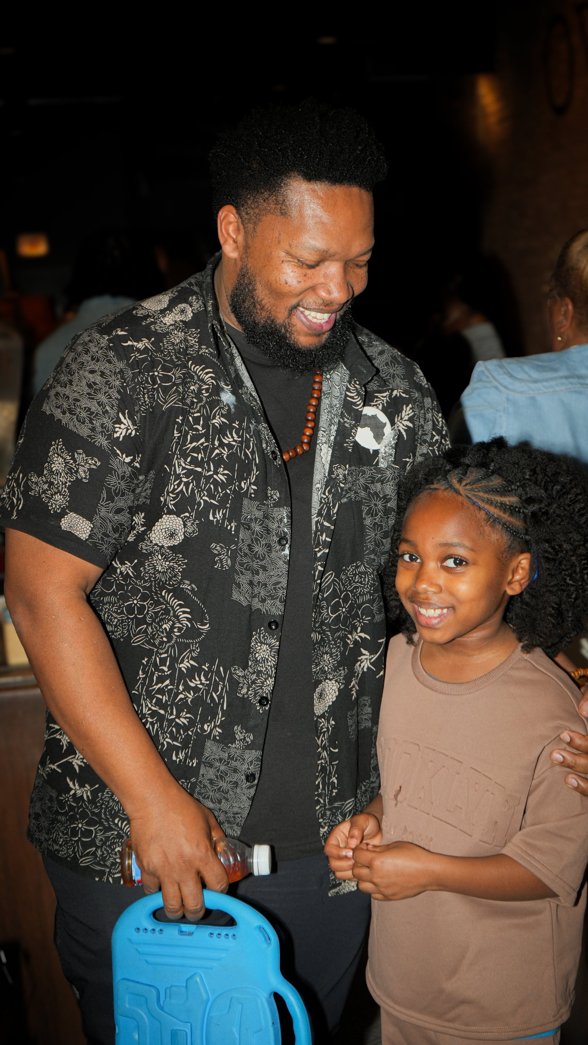 A man with a beard and a young girl smiling together at an indoor event, the man holding a blue toy and the girl wearing a brown T-shirt.