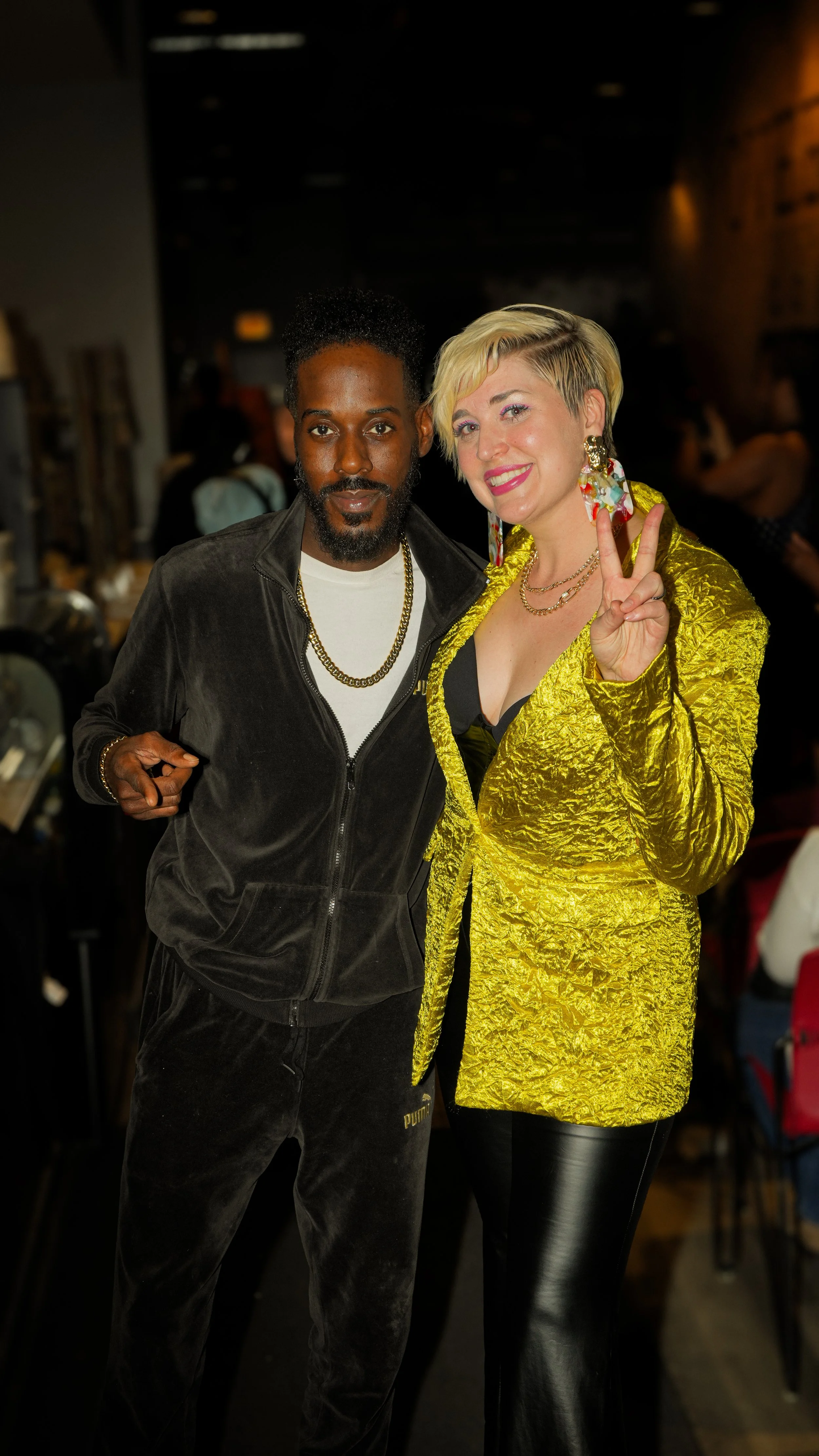 Two people posing together inside a dimly lit venue. The person on the left is a dark-skinned man with curly hair, wearing a black zip-up jacket and gold jewelry. The person on the right is a light-skinned woman with short, blonde hair, wearing a shiny yellow blazer, earrings, and makeup, making a peace sign with her right hand.