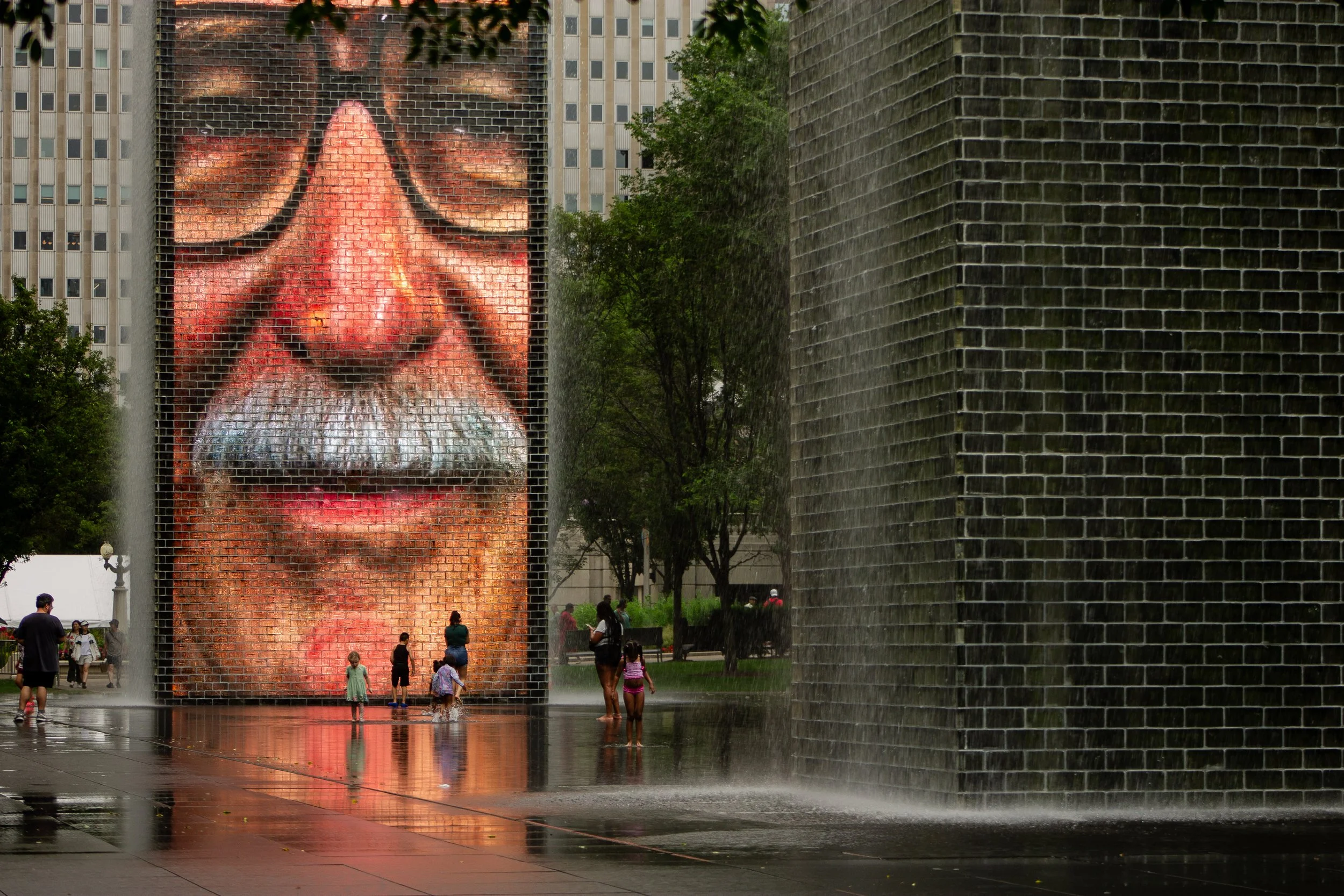A large outdoor mural on a building wall depicts a man's face with glasses, nose, and lips. There are trees and water fountains nearby with people walking and standing around.