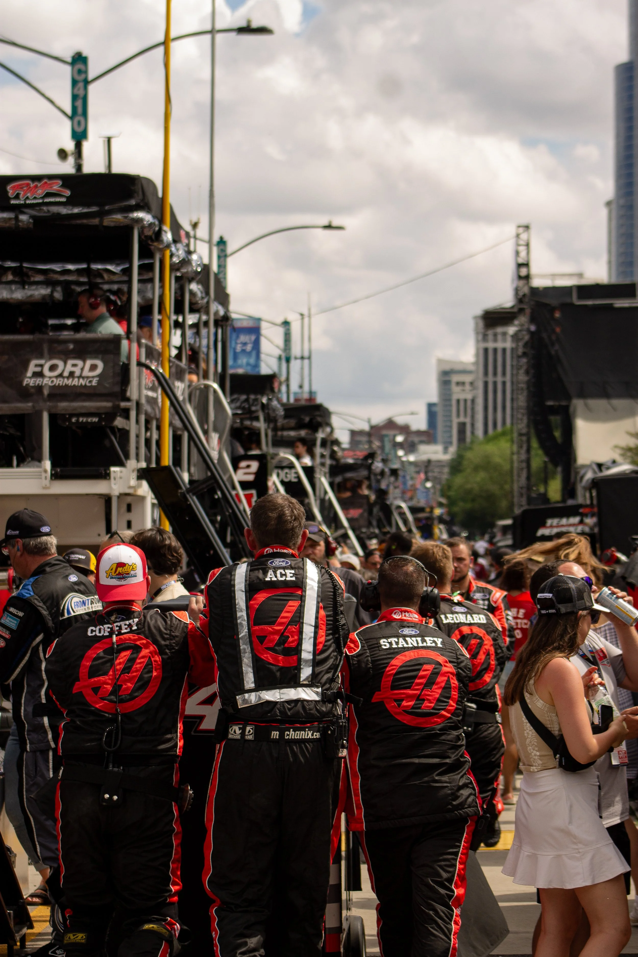 Race car drivers in black racing suits with red accents and sponsor logos, gathered in a pit lane area, with team members and onlookers nearby, and trailers and buildings in the background on a partly cloudy day.