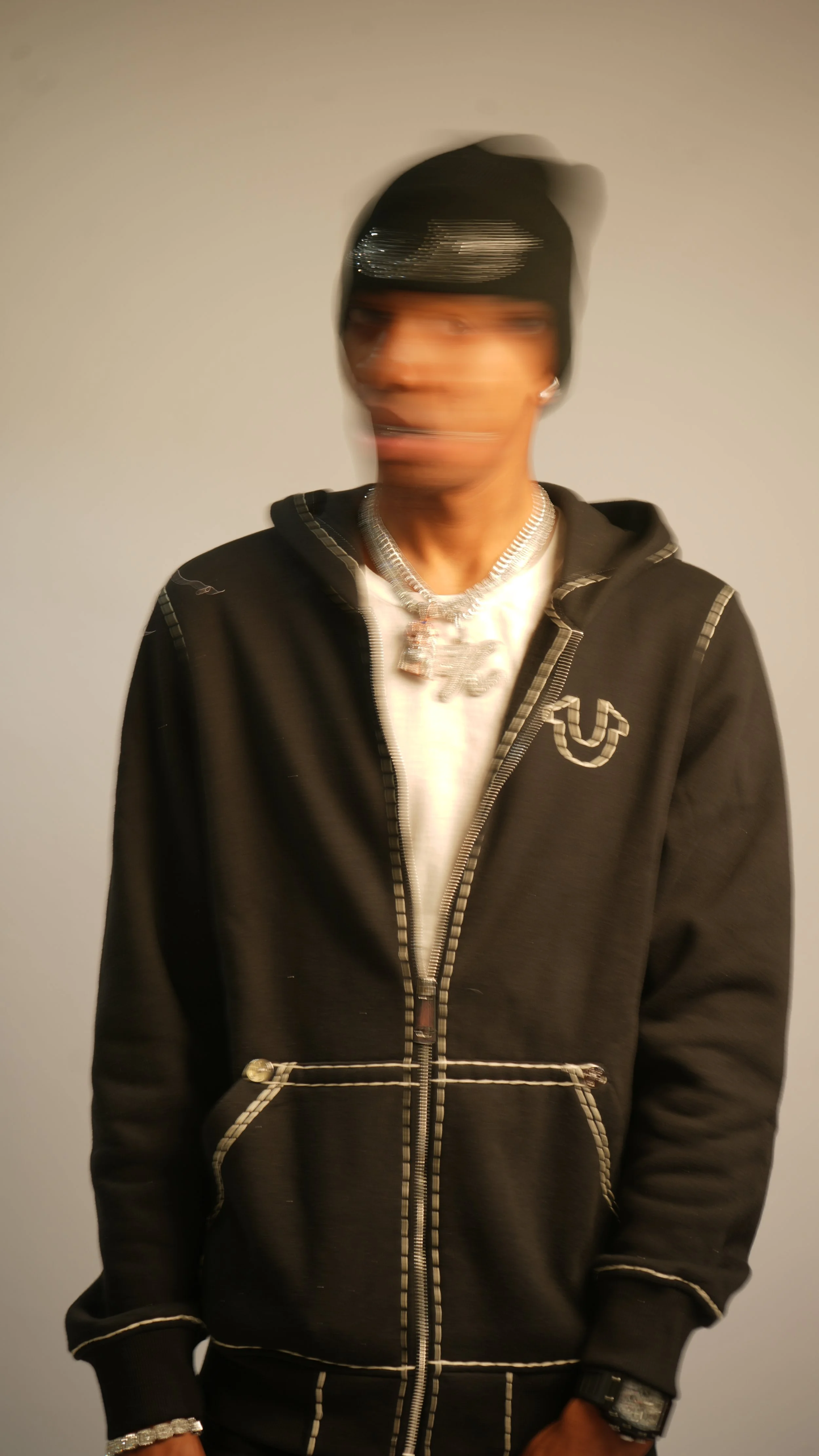 A person wearing a black hoodie with white stitching, a black beanie, silver jewelry, and a white shirt, standing against a plain wall. The image is blurry with motion.