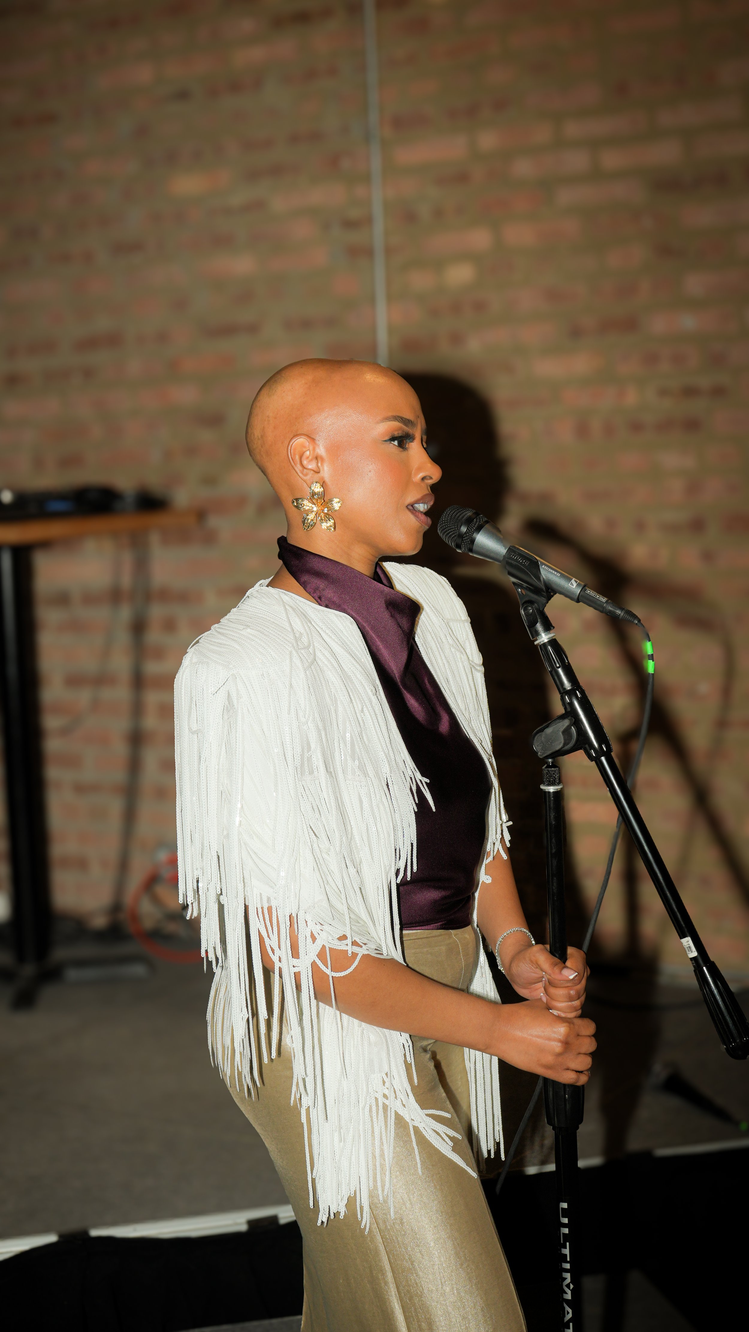 A woman with a shaved head singing into a microphone at an indoor event with a brick wall background.