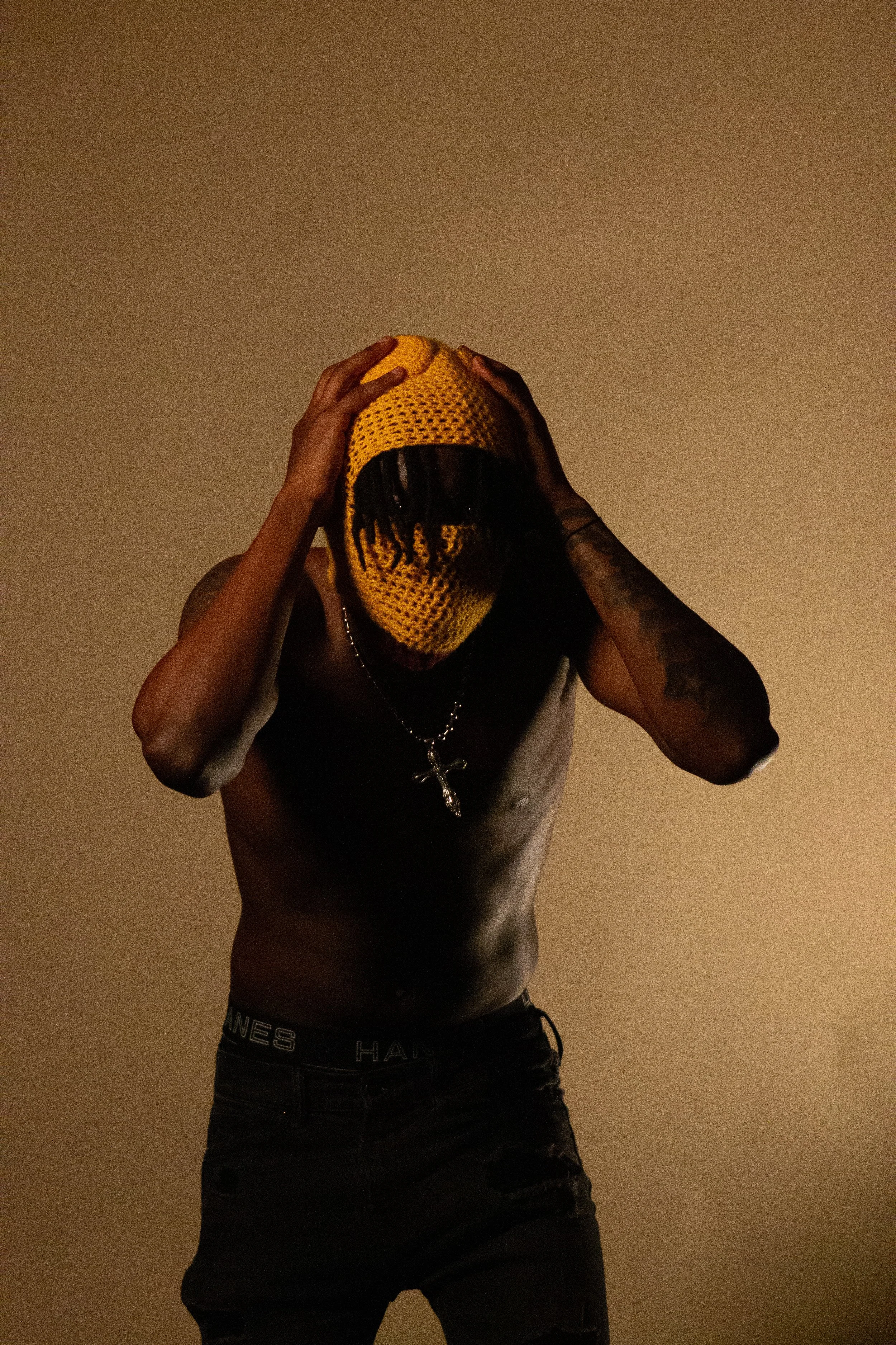 Person wearing a yellow knitted ski mask holding their head with both hands, standing against a plain wall, dressed in black pants and a necklace.