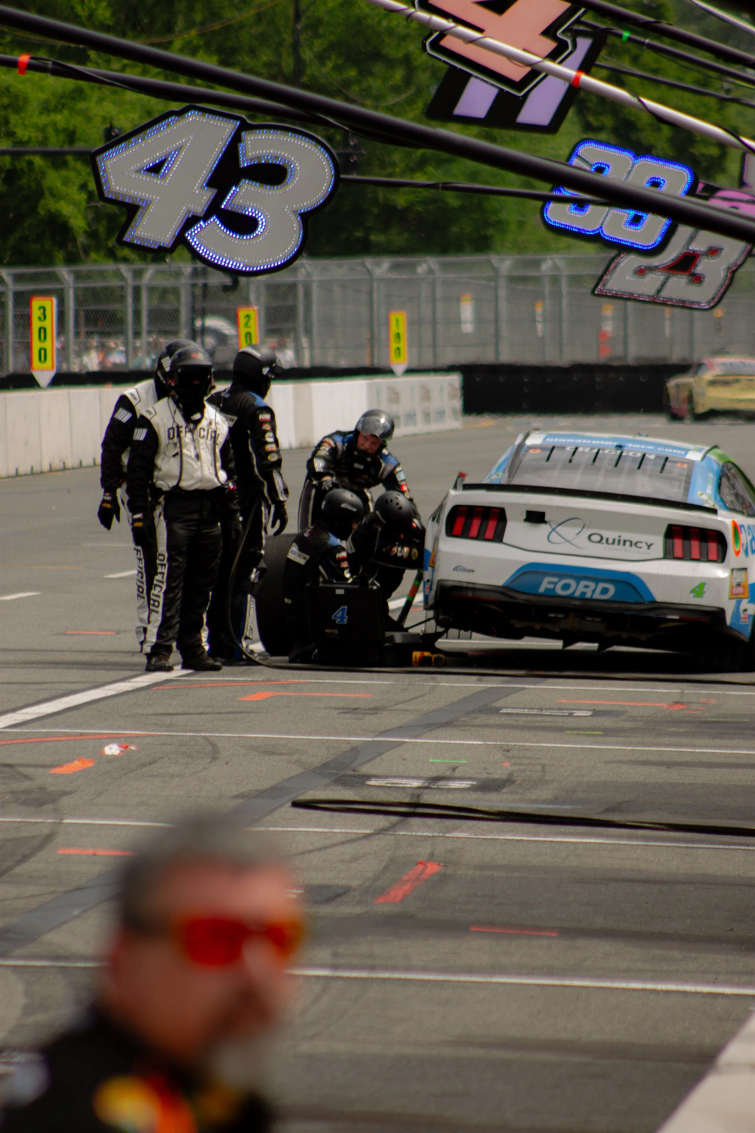 Race car pit stop scene showing a group of mechanics working on a white and blue Ford race car, with race flags showing the number 43 and 88 hanging above. Several people in racing suits and helmets are near the car.