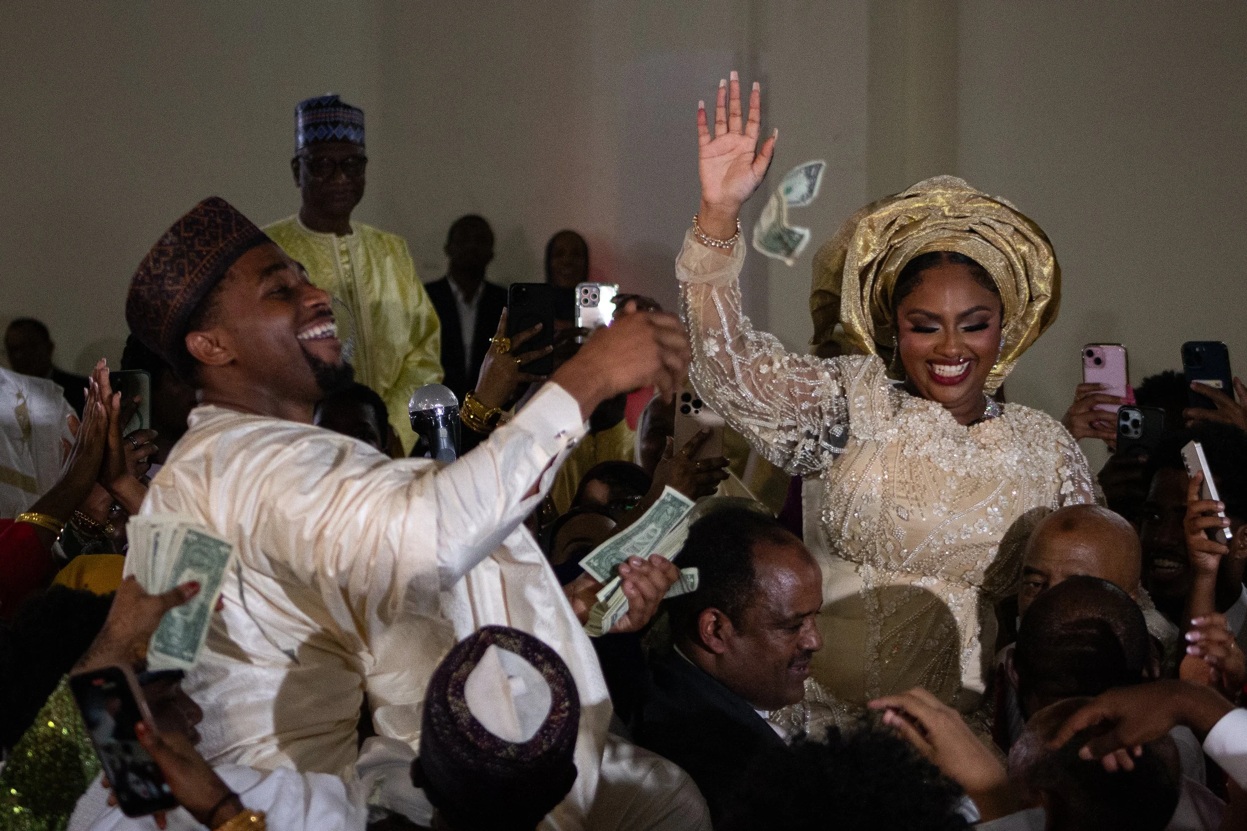 A woman dressed in an elegant traditional dress with a large headwrap is smiling and raising her hand, surrounded by people taking photos and throwing money in celebration.